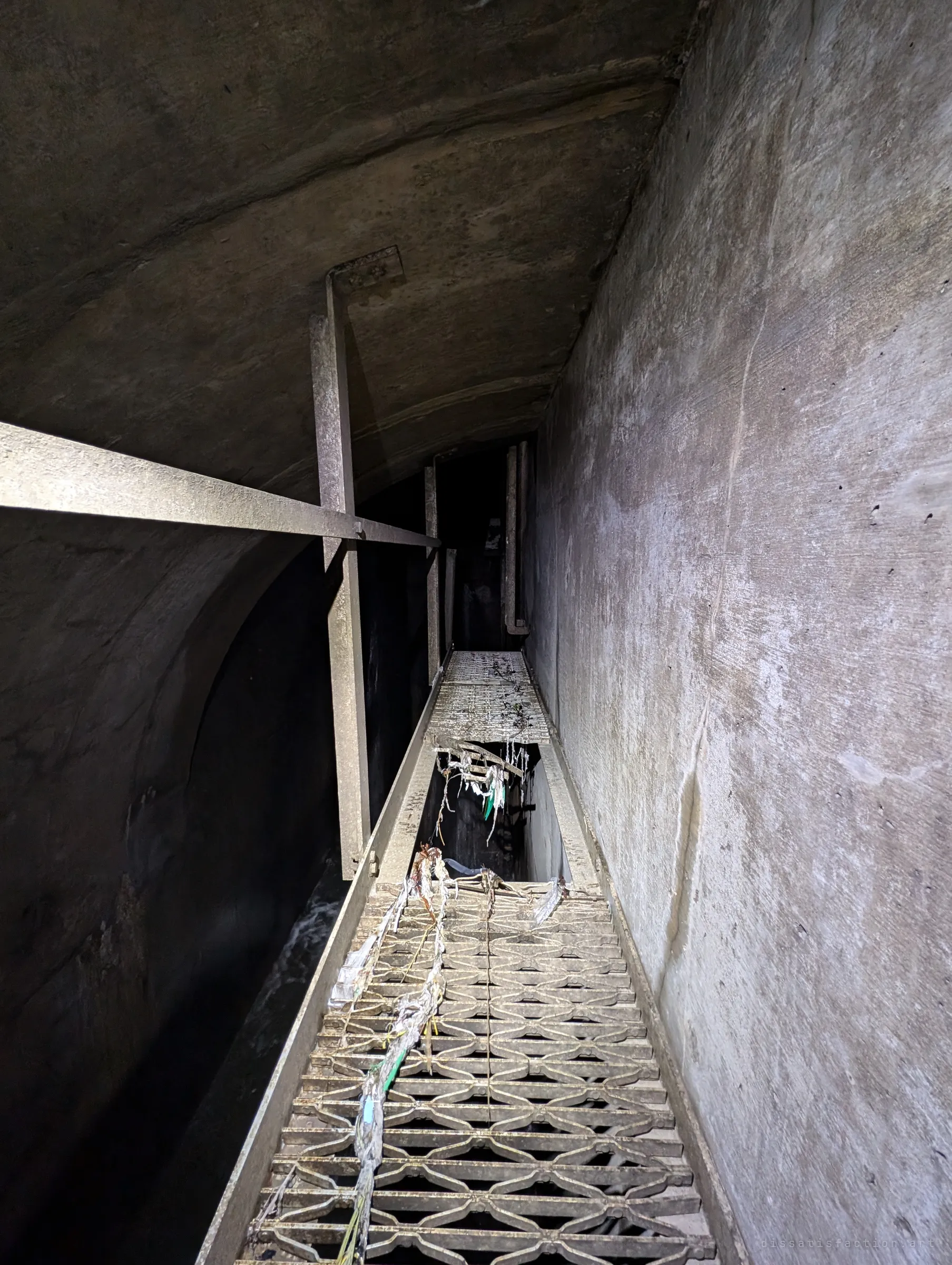 Looking down a narrow grated catwalk high above the flow, with a missing section opening straight to the water below and rag debris snagged along the steel.