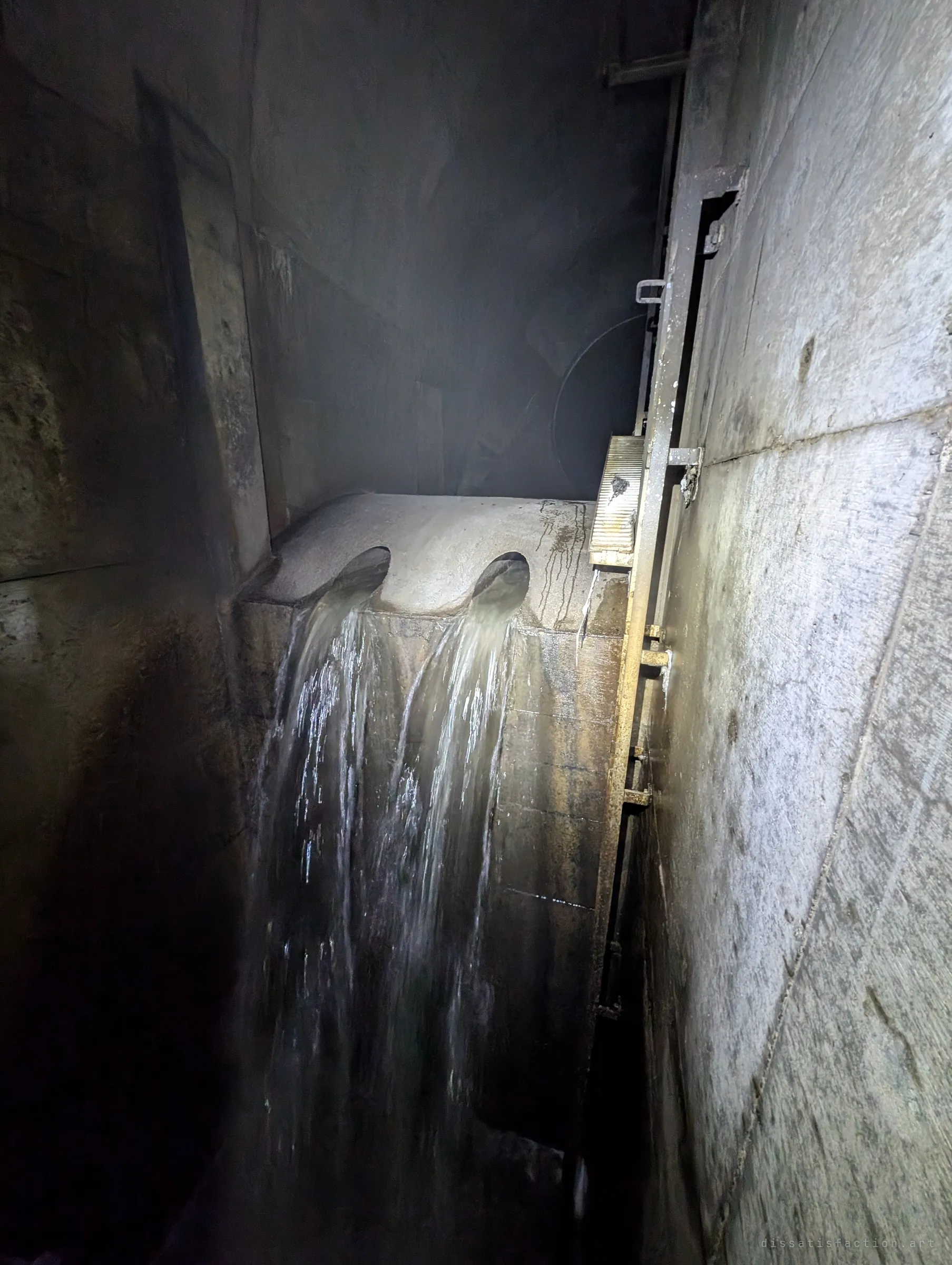 Water pours through two round openings in a concrete wall into a deep shaft, beside a steel gate frame lit by flashlight in the misty dark.