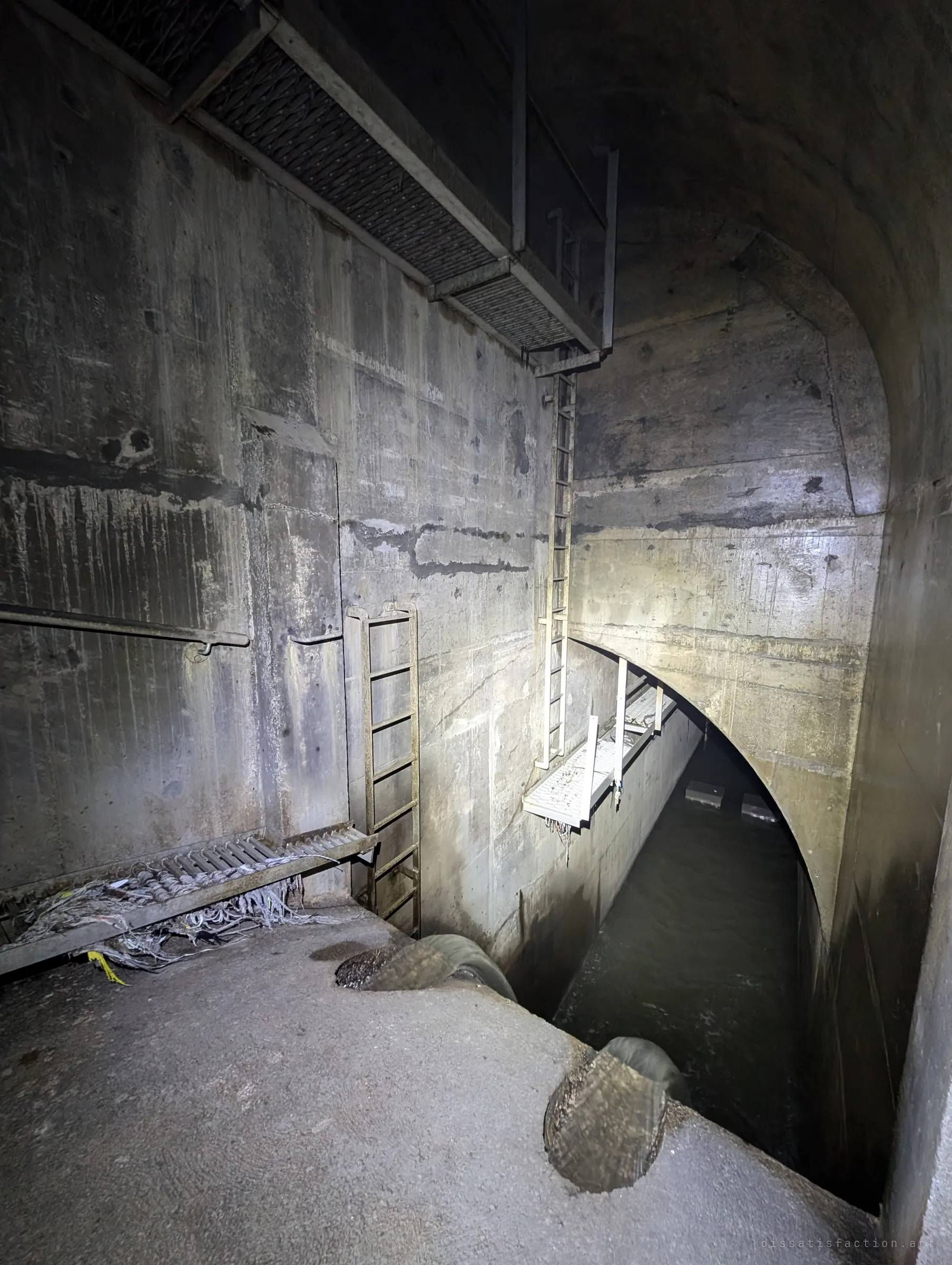 Looking down from a concrete ledge into a deep storm-drain chamber, with steel ladders, grated catwalks and a dark channel running off under a wide arch. The ladders and walkways show how the chamber is built for maintenance access.