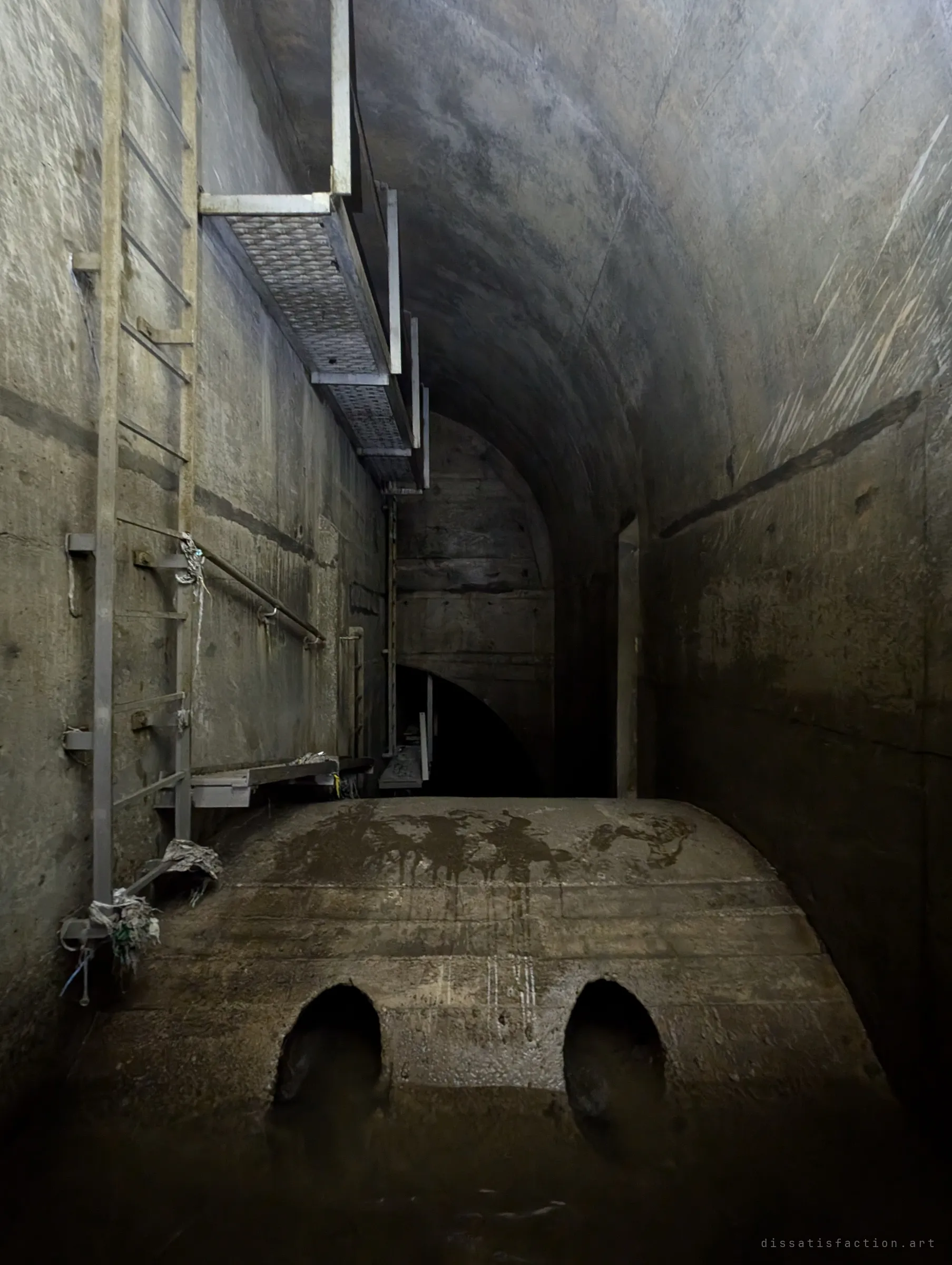 A tall vaulted drain chamber where a rounded concrete weir with two arched openings holds back the water, below a catwalk and ladder bolted to the wall. Debris snagged on the ladder hints at how high the water rises.