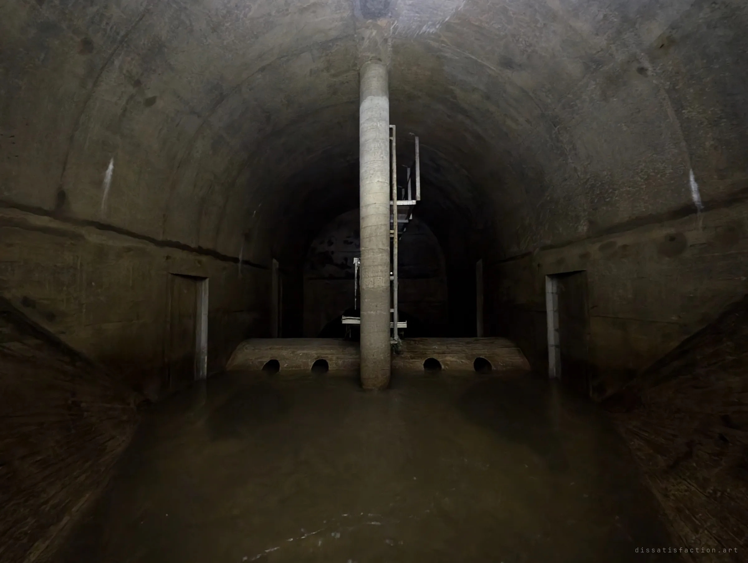A single round concrete column stands in the middle of a wide arched chamber, in front of a low weir pierced by four small openings. Still brown water fills the floor and the tunnel continues into darkness beyond.