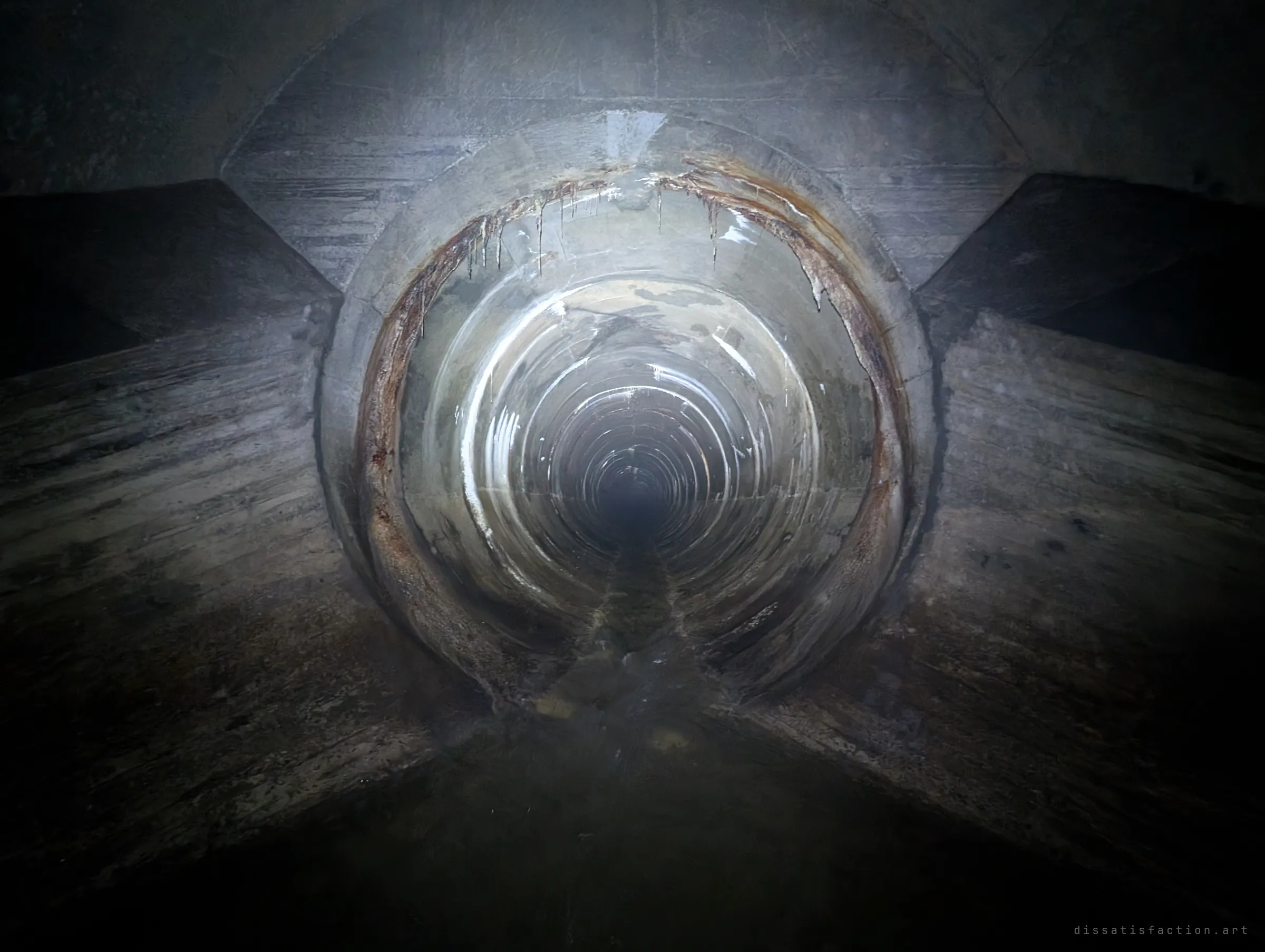 A round pipe opening in the chamber wall, rimmed with rust-stained drips, recedes into a long glowing tunnel of concentric rings. The flashlight turns the pipe into a near-perfect vanishing point.