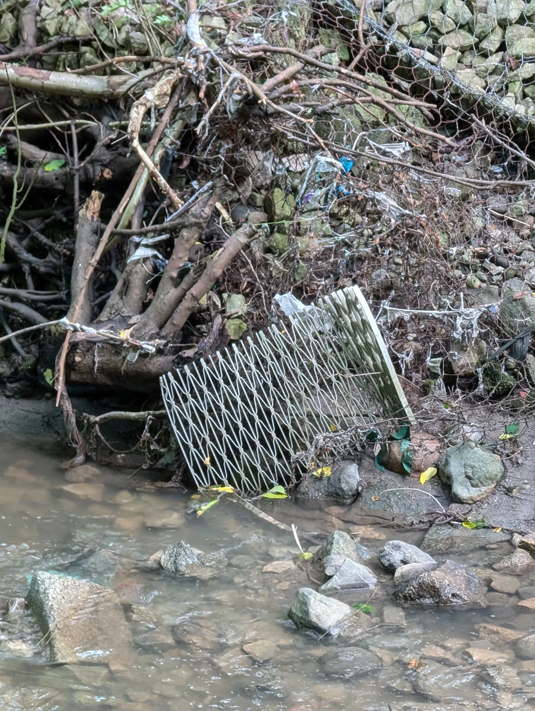 A bent metal grate lies tangled in branches, chicken wire and shredded plastic on a creek bank. Storm debris jammed against the rocks shows the force of the water that passes through.