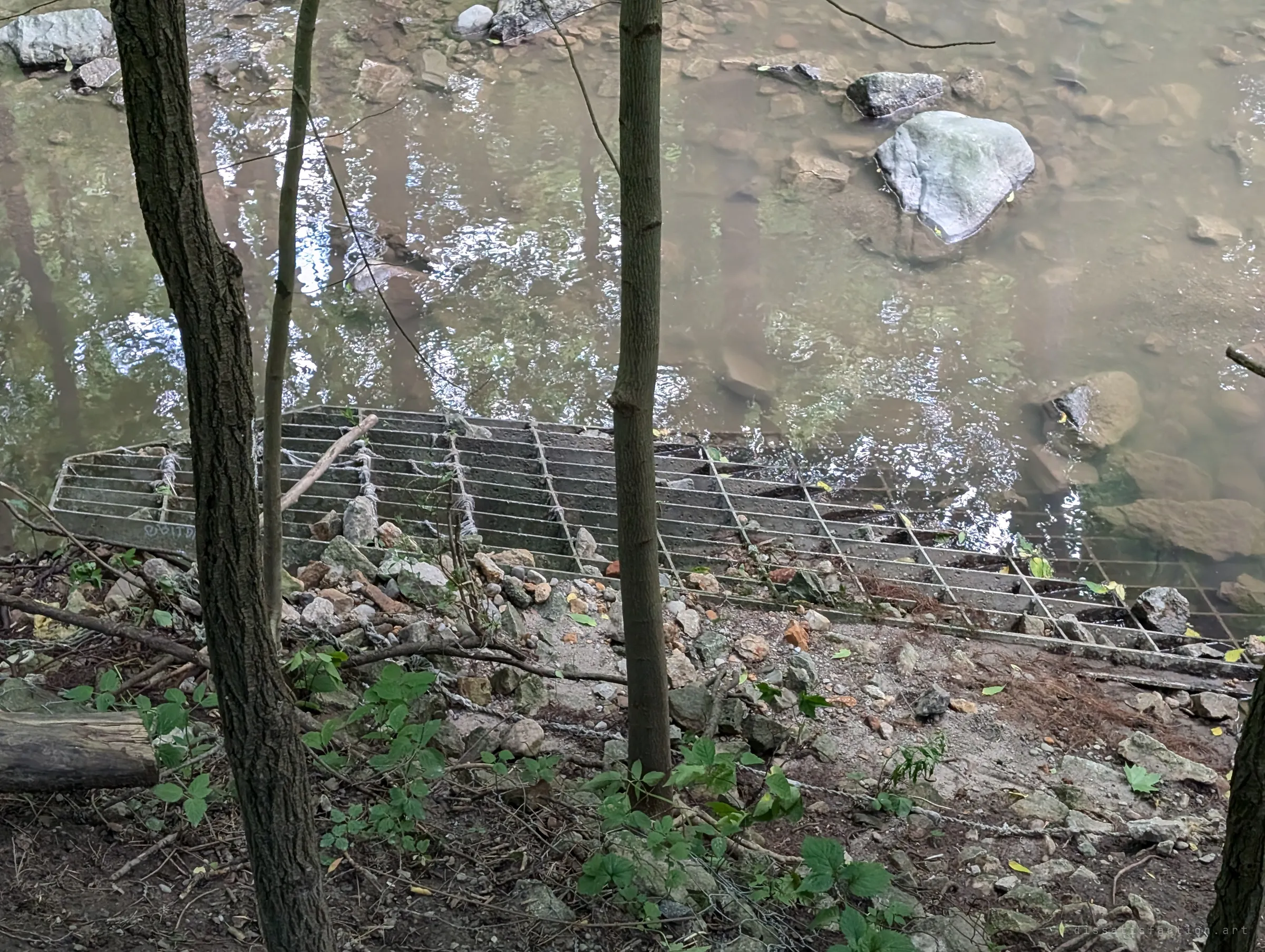 Seen from the wooded bank, a large steel grate lies twisted and half buried in rubble at the edge of a shallow creek. It was torn loose and dragged here by floodwater.