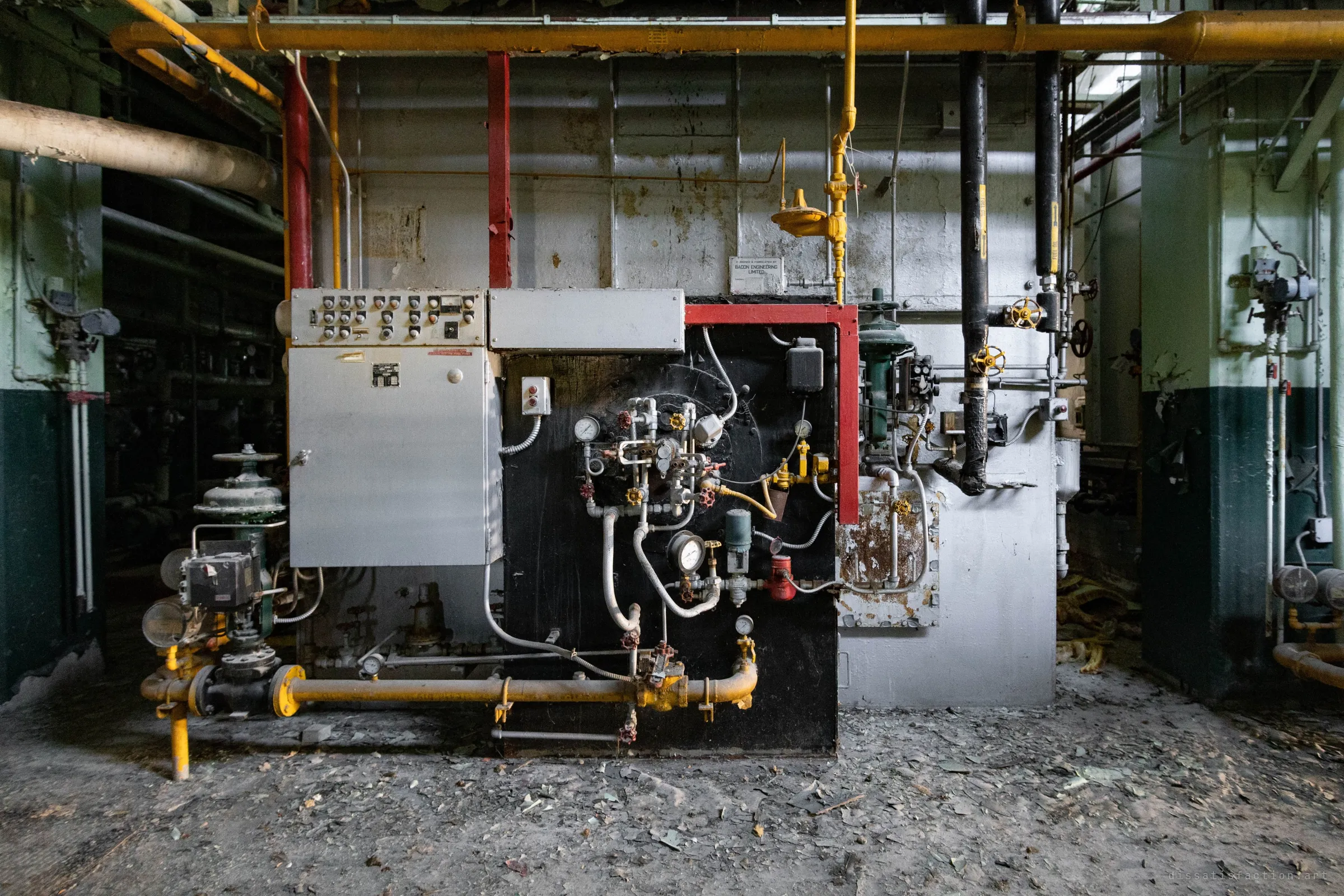 Front of a gas-fired boiler: a grey control cabinet, a black burner plate crowded with gauges and valves, yellow gas lines and red framing, debris across the floor.