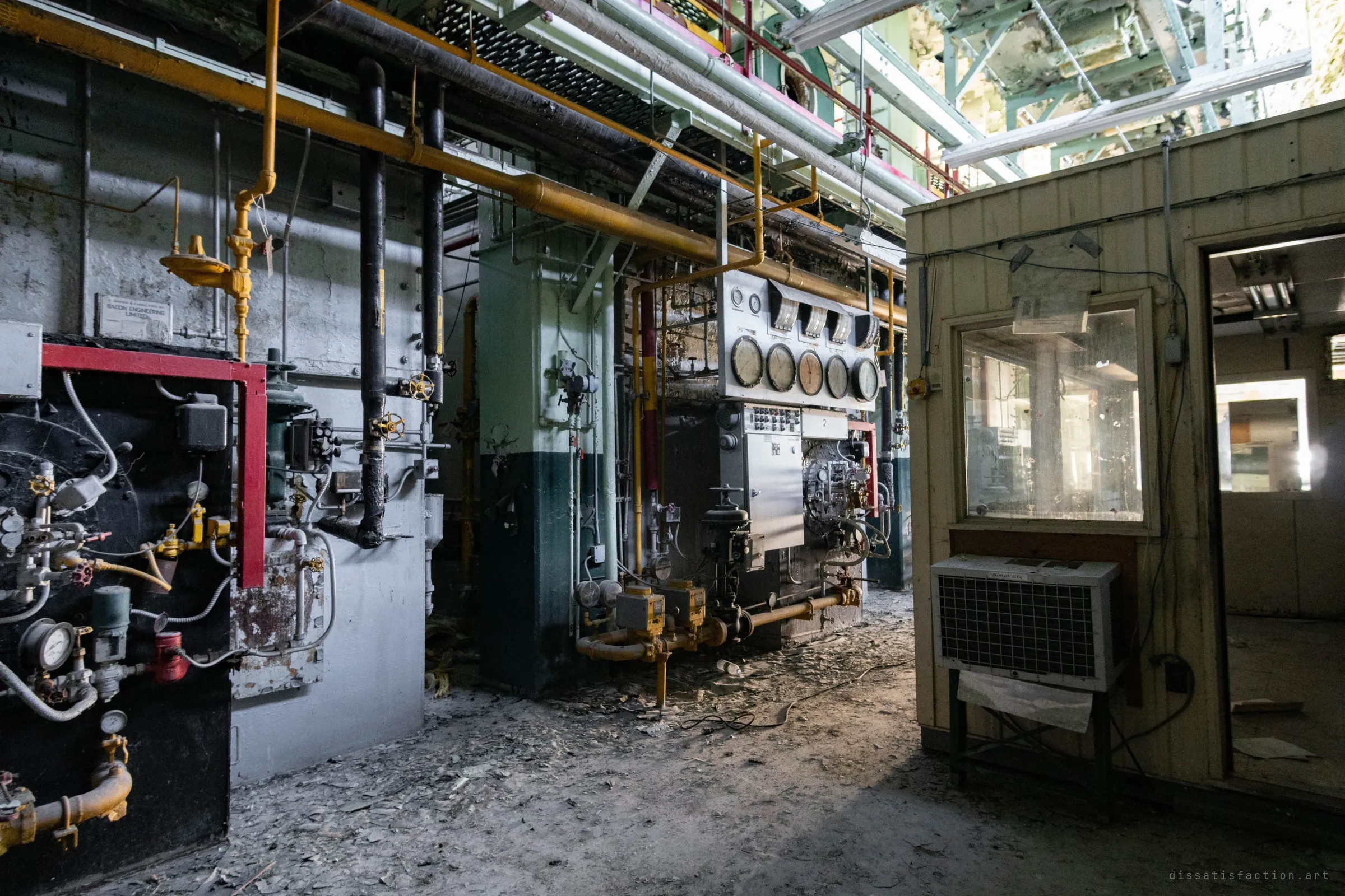 A row of boilers seen side-on, big dial gauges on a control panel, and a small wooden operator's booth with a window air conditioner under a torn-open ceiling.