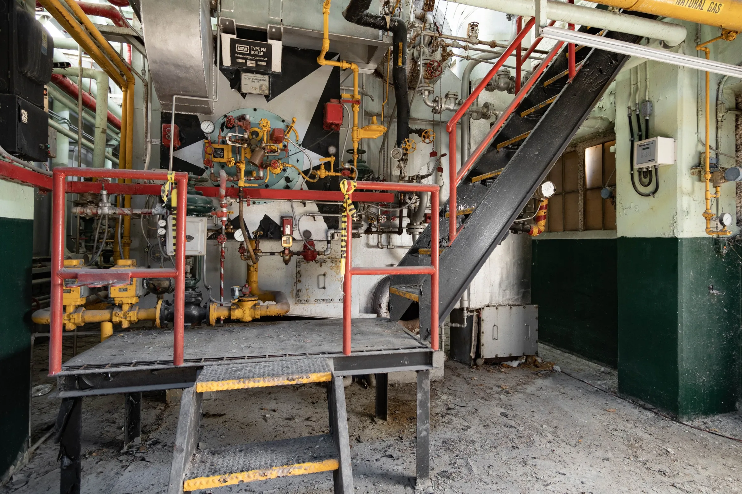 A boiler front painted with black and white starburst panels behind a red-railed step platform, caution tape hanging loose and a black stair climbing to the right.