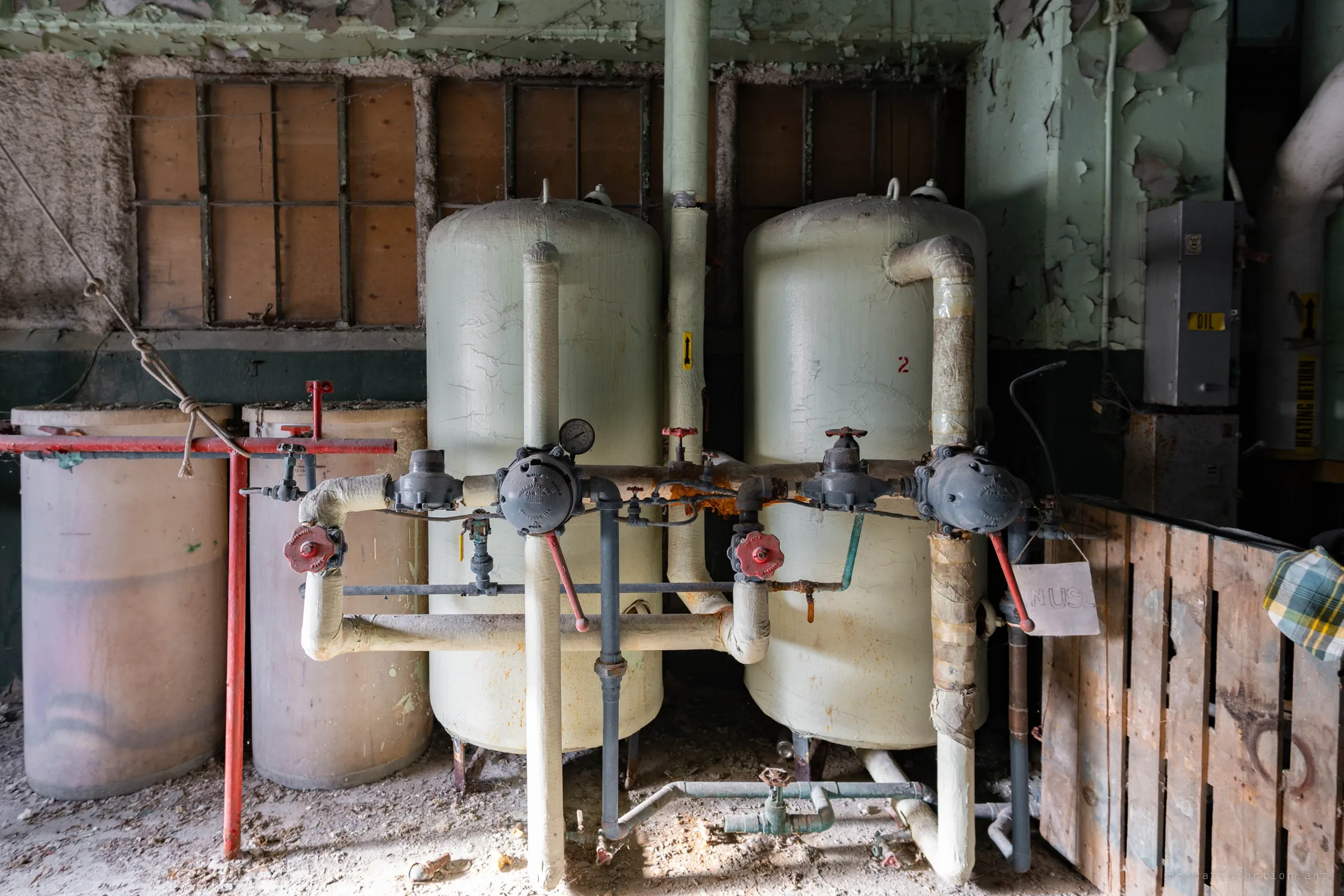 Two pale water-treatment tanks with cracked insulation and grey valve bodies, between old drums and a pallet, in front of boarded windows and peeling green paint.