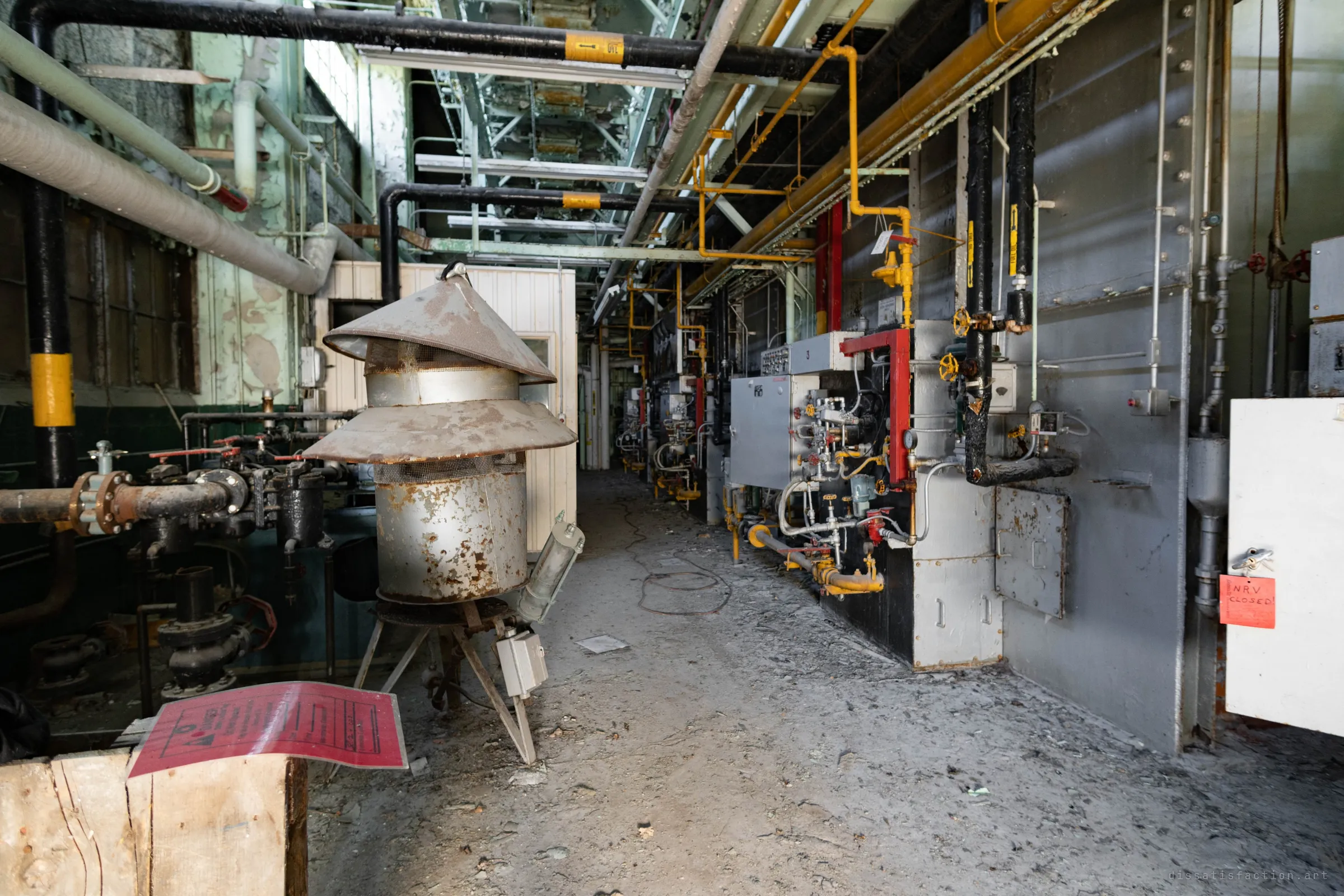 A long aisle past a rusted conical vent cap, with the boiler fronts lined up on the right and a handwritten red tag hanging off a cabinet.
