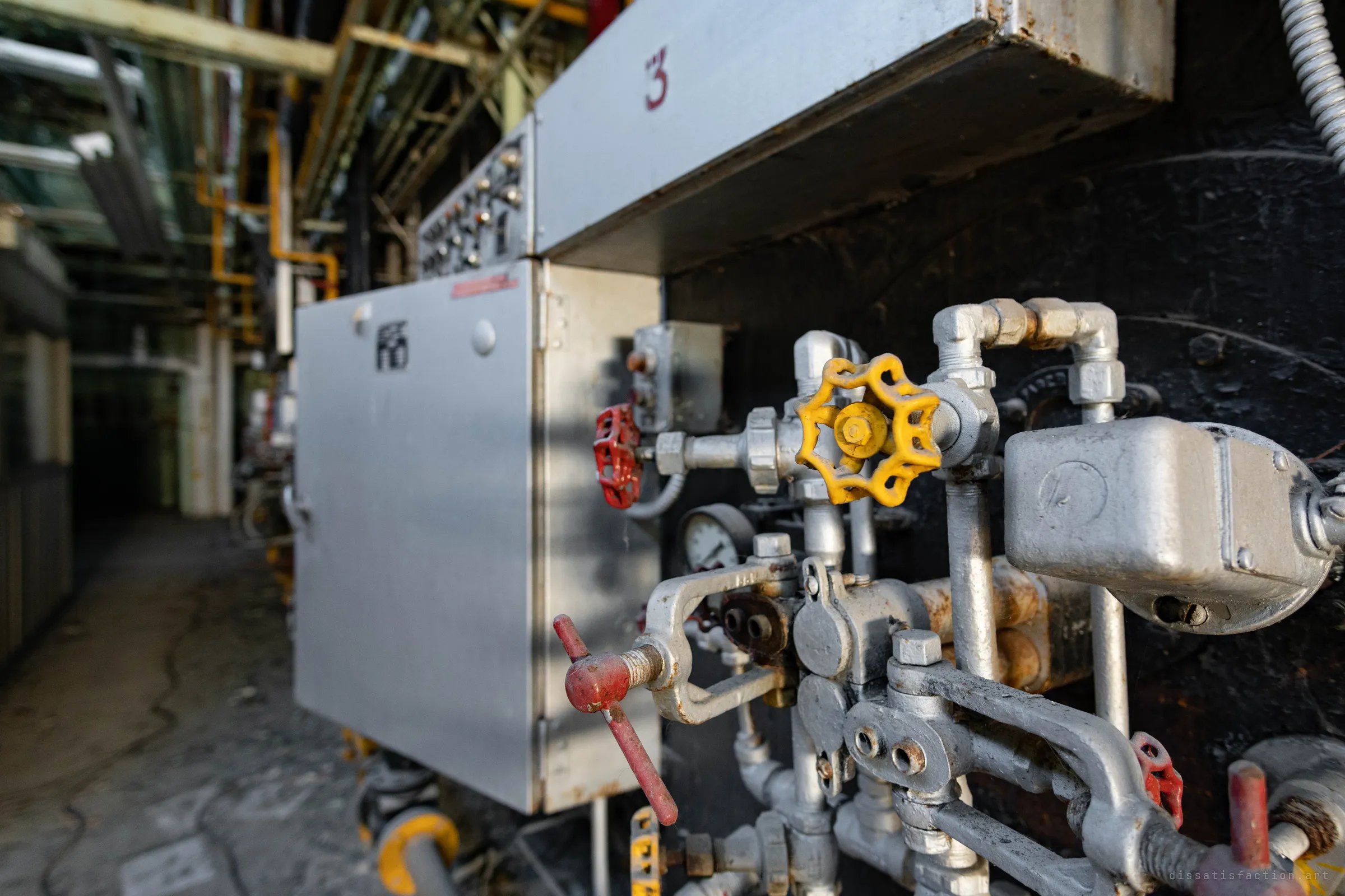 Close-up of a burner's valve cluster, yellow and red handwheels sharp in front, with the numbered control cabinet and the long boiler aisle blurred behind.
