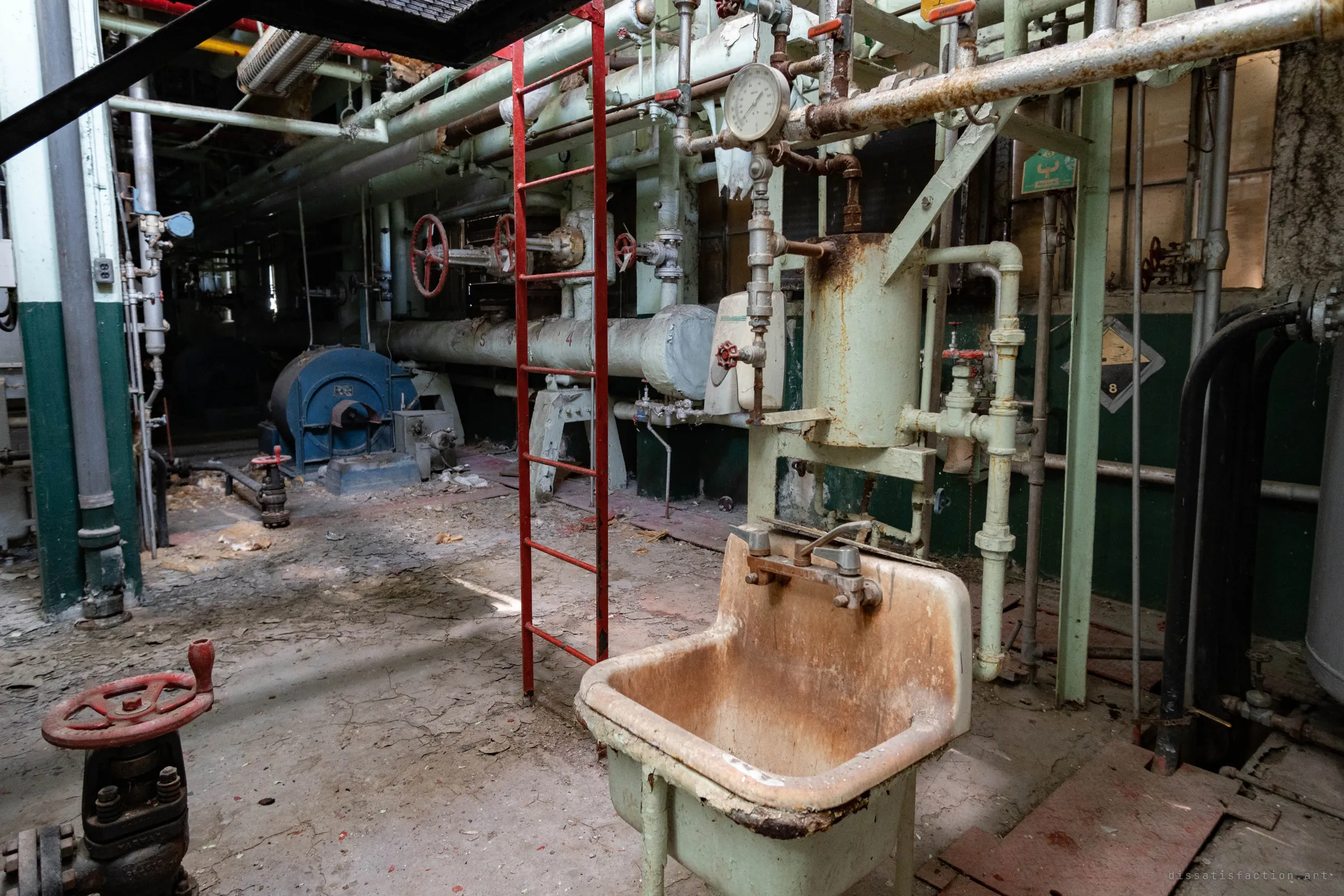 A stained utility sink under a rusted water heater, with a red ladder and red handwheels beside it and a blue fan housing along the back wall.
