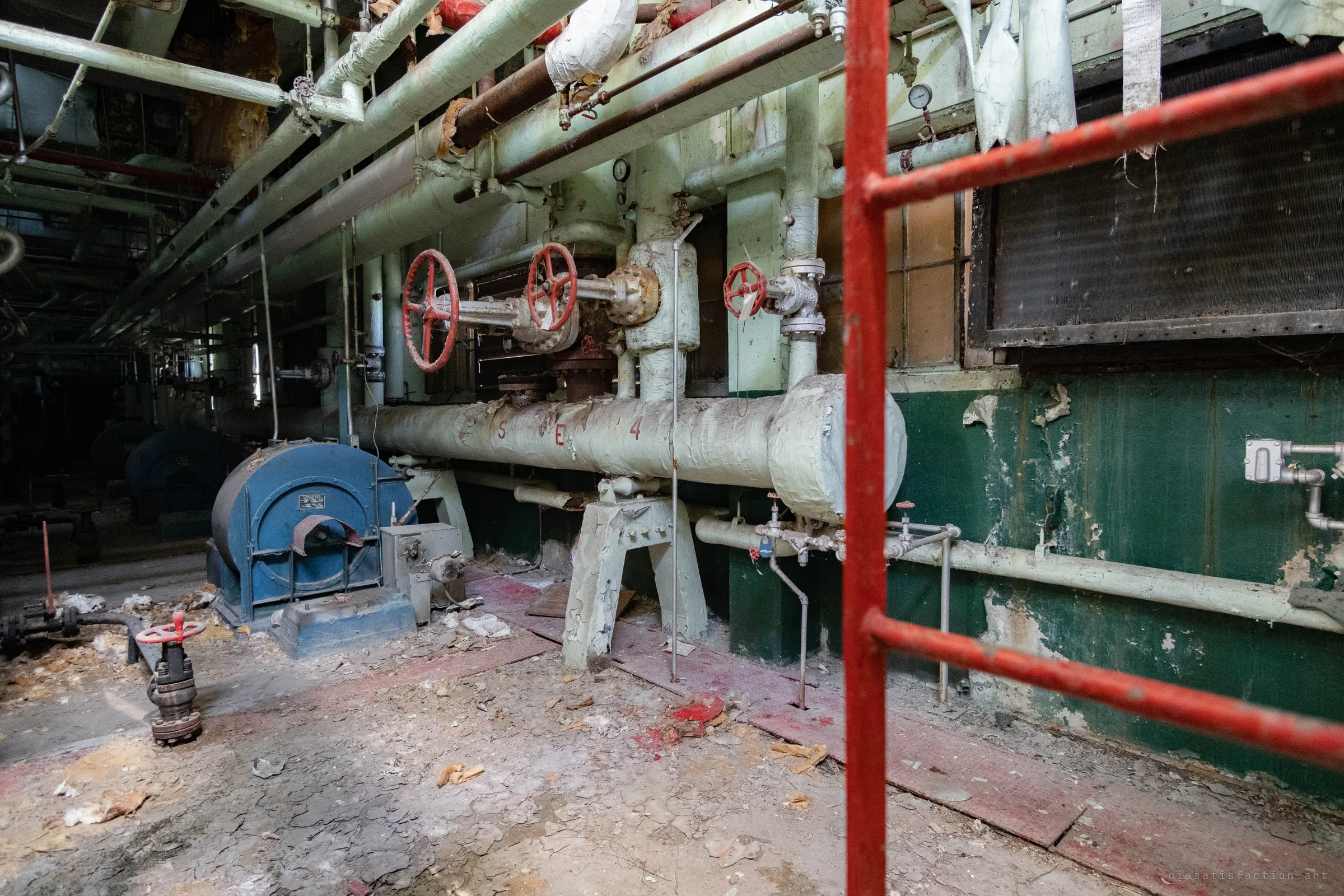Seen through a red ladder: a lagged steam header marked with painted letters and a number, red gate valves above and a blue fan motor on the littered floor.