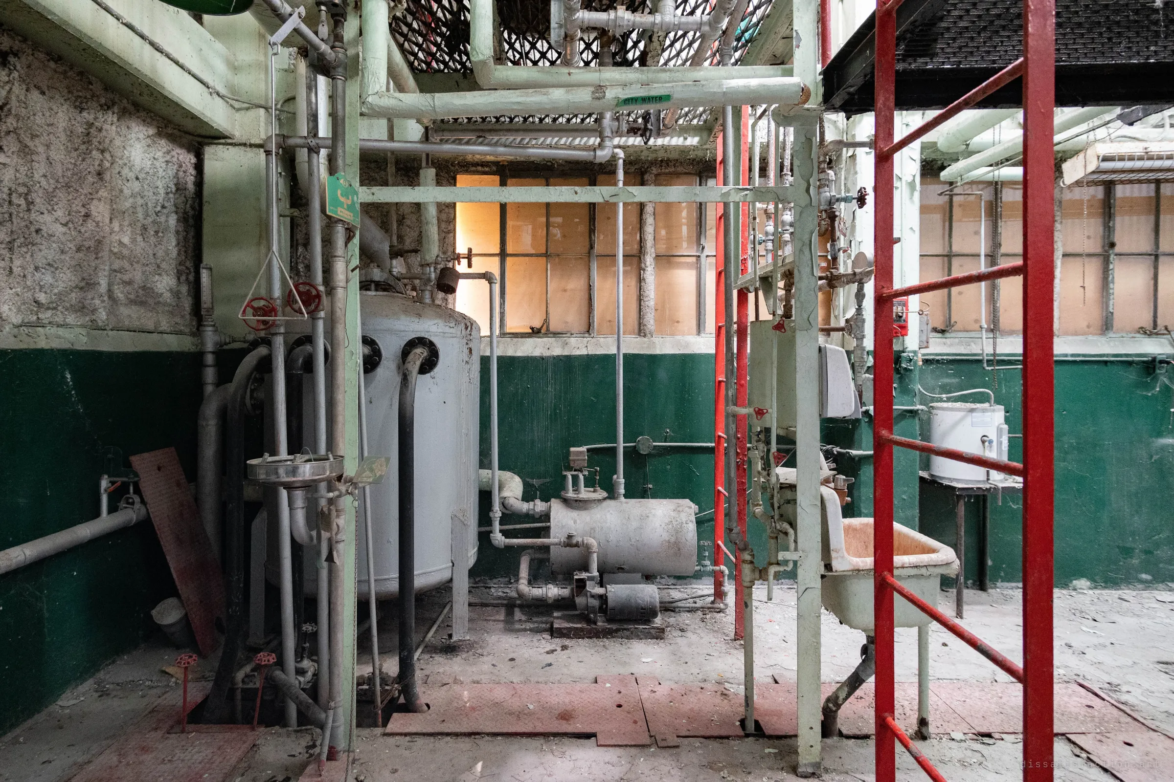 A symmetrical pump corner: a grey pressure tank and a small pump against a two-tone green wall, amber light through boarded windows, red ladders framing it.