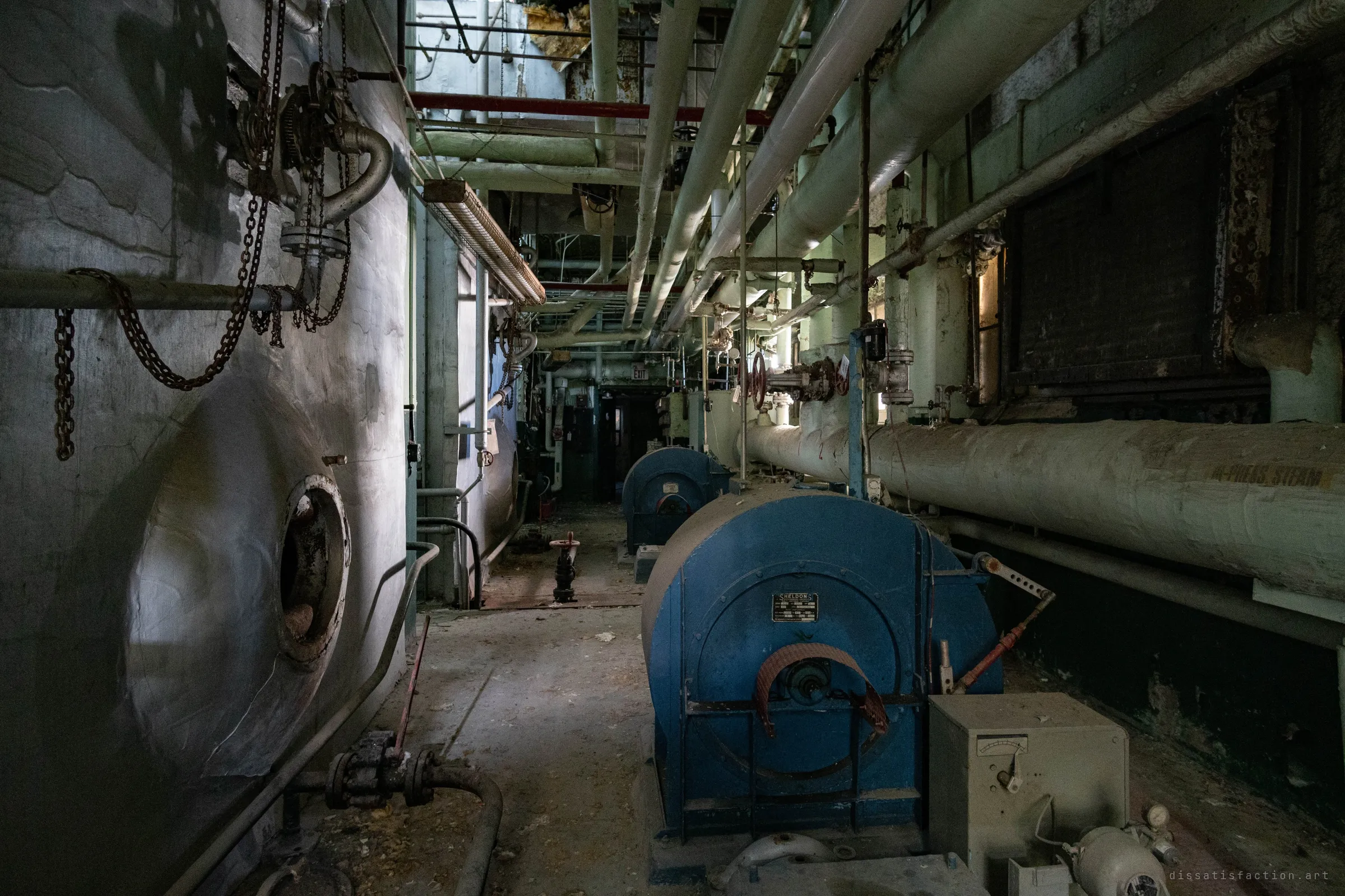 A dark corridor between a boiler shell and a row of blue draft fans, chains hanging in front and insulated steam lines running off into the gloom.