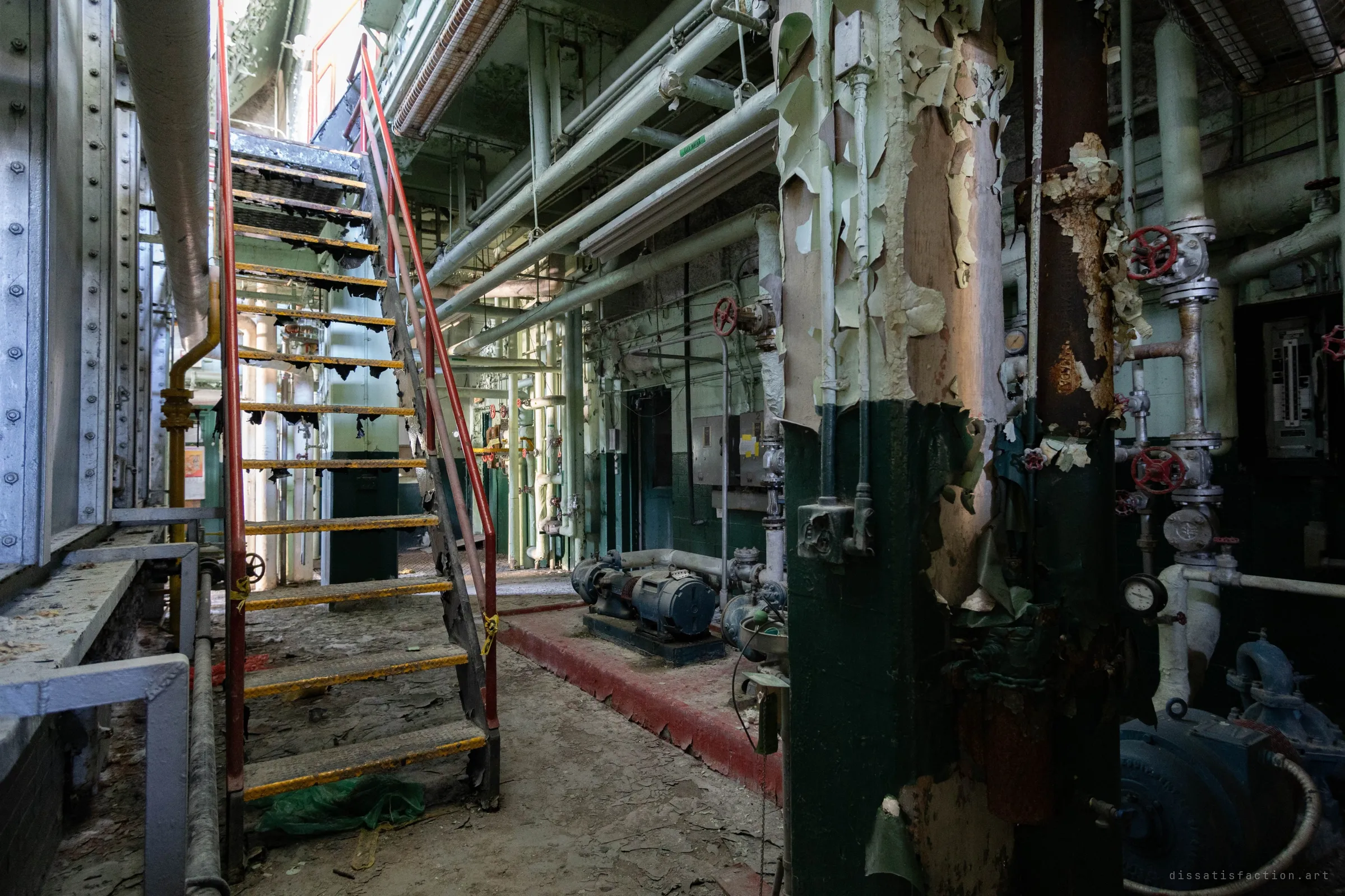 A red-railed stair climbs past a column shedding sheets of paint, with pumps and valves along the aisle and daylight falling in from above.
