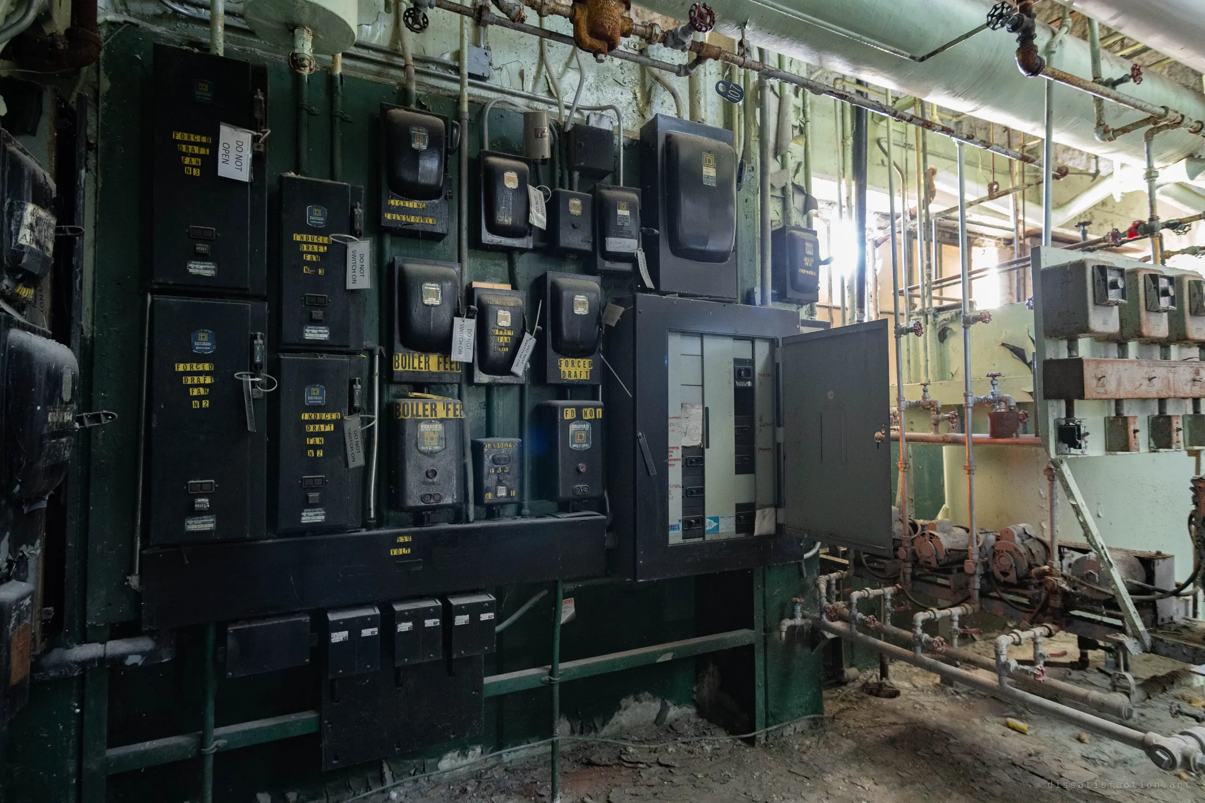 A wall of black motor starters with stencilled fan and feed labels and "do not switch on" tags, beside an open breaker panel and a run of rusty pipes.