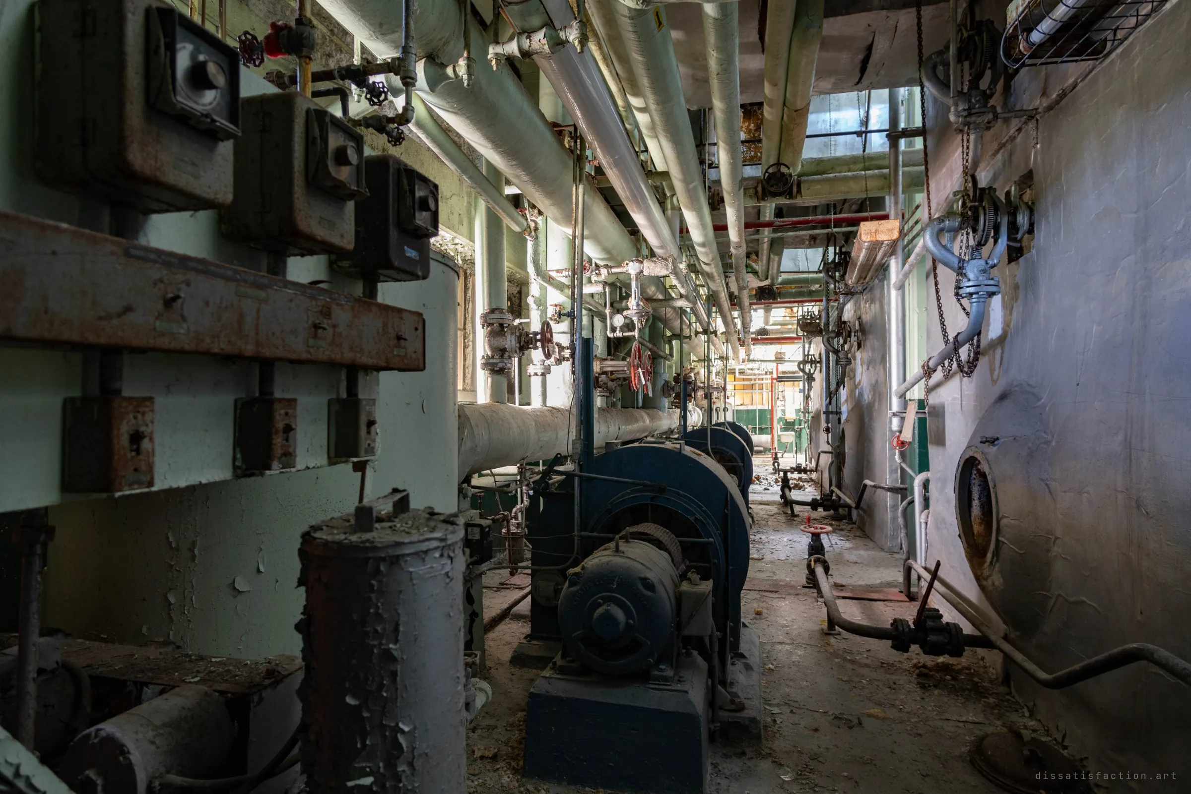 A narrow gap between a boiler shell and a line of blue fans, rusted starter boxes on the left and pipes packed overhead toward a bright far end.