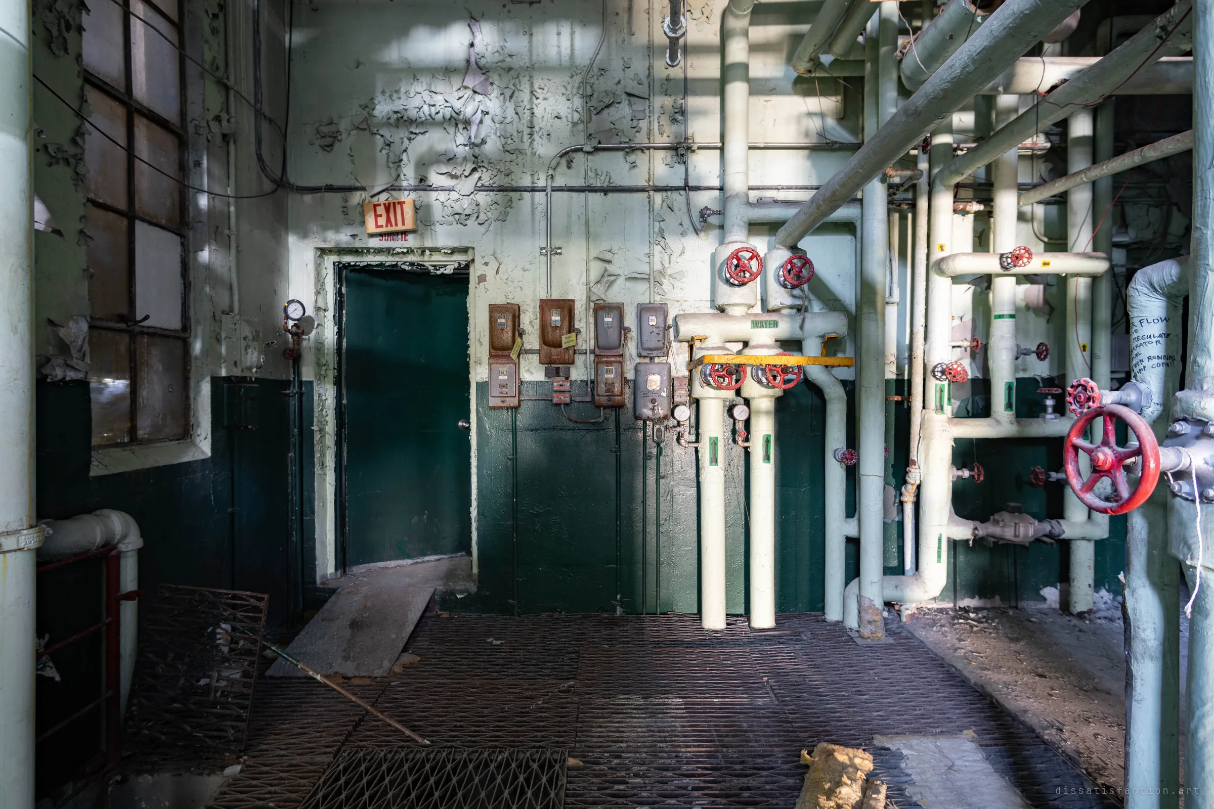 A green steel door under an exit sign, a row of old switch boxes beside it and white water lines with red handwheels, one pipe covered in handwritten notes.