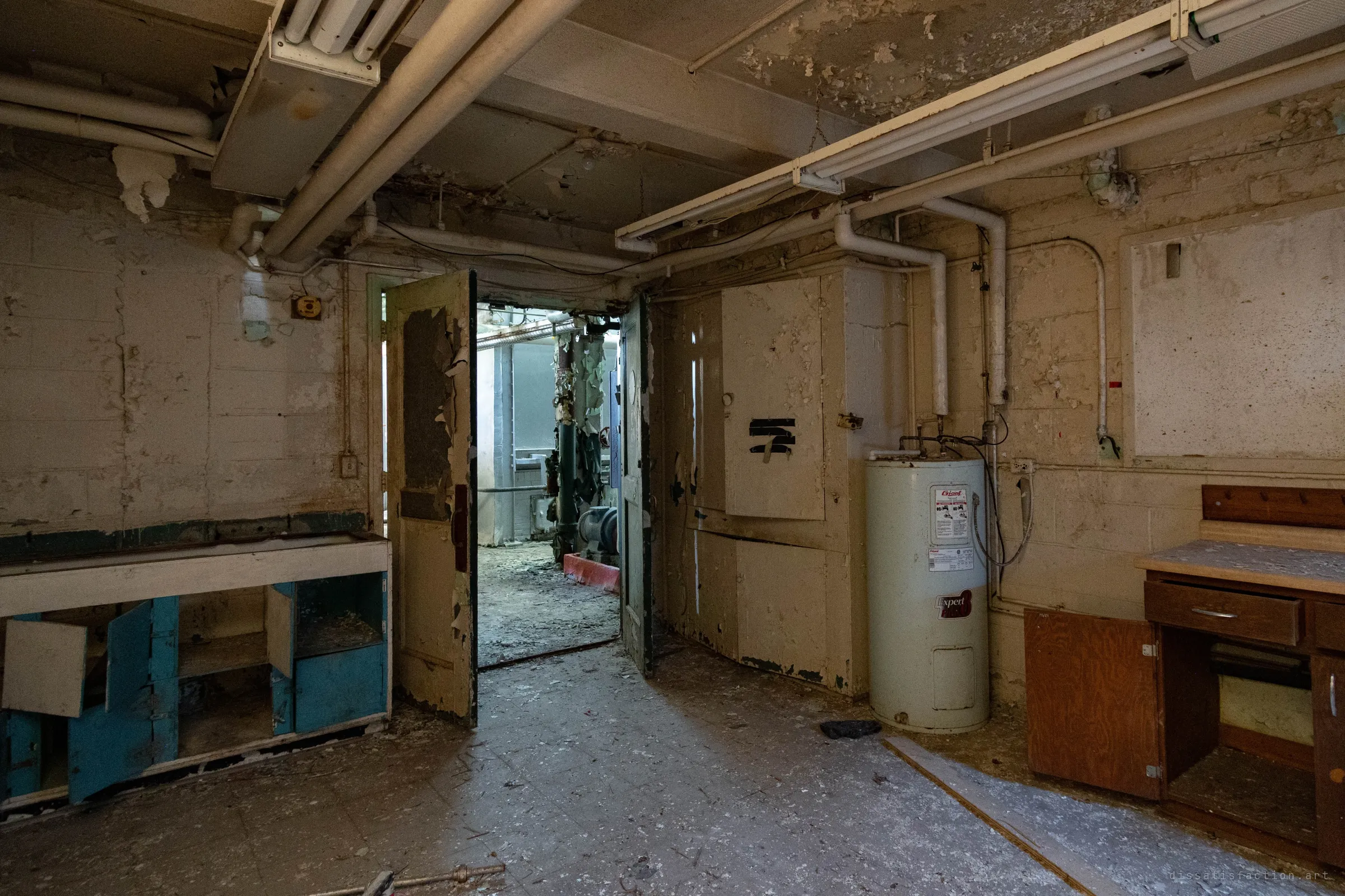 A basement workroom with blue lockers, a water heater and battered cabinets, its double doors open onto the lit pump room beyond.