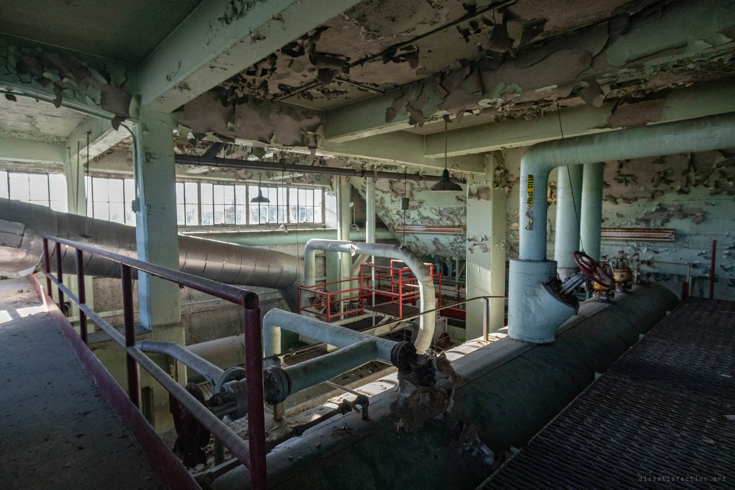 Looking down from the upper walkway to red-railed platforms below, a huge insulated duct slanting past a row of windows, paint curling off the ceiling.