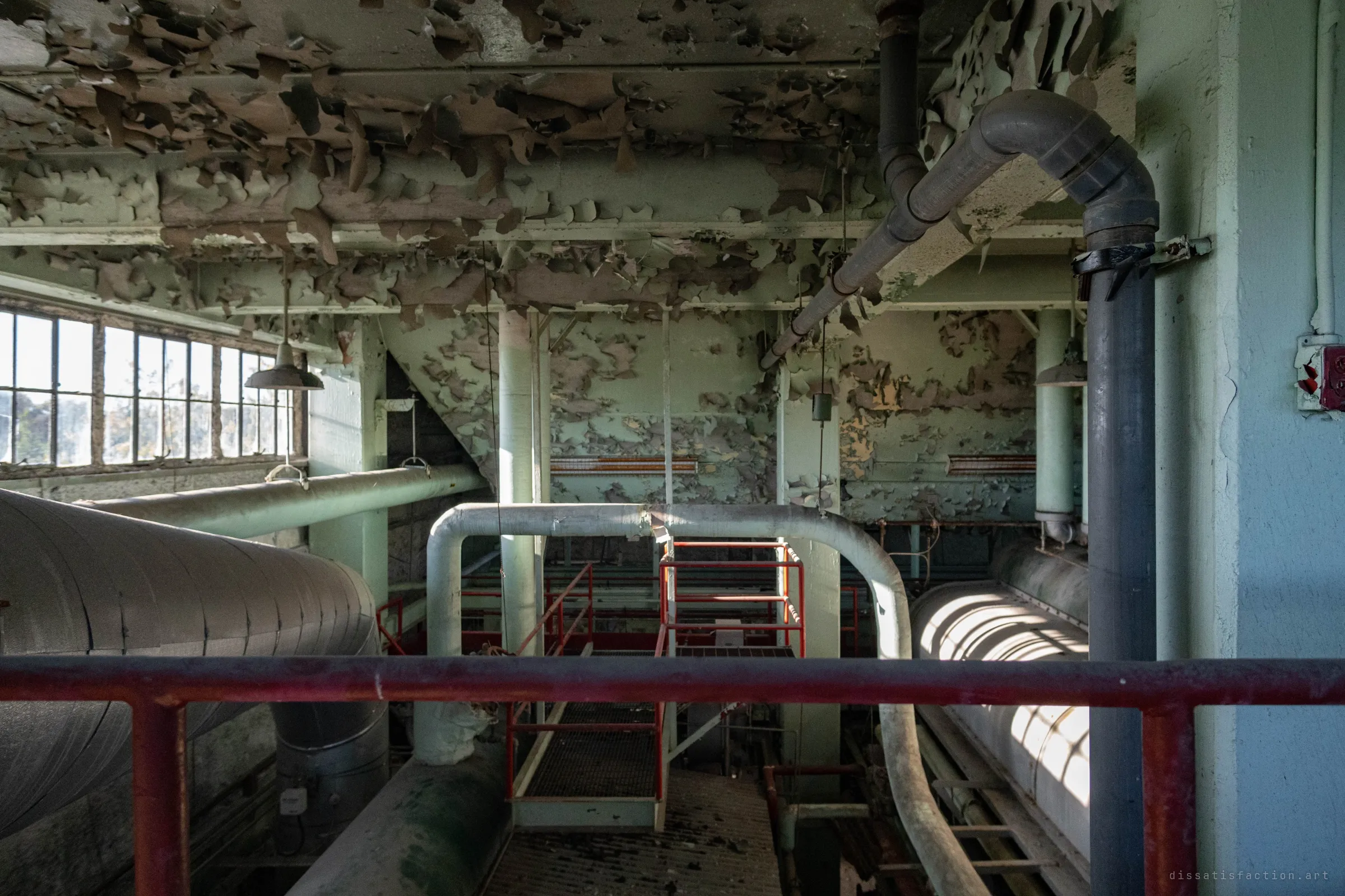 From behind a red railing, a pipe arches over a caged platform in the pit below, lit from side windows under a ceiling of peeling paint.