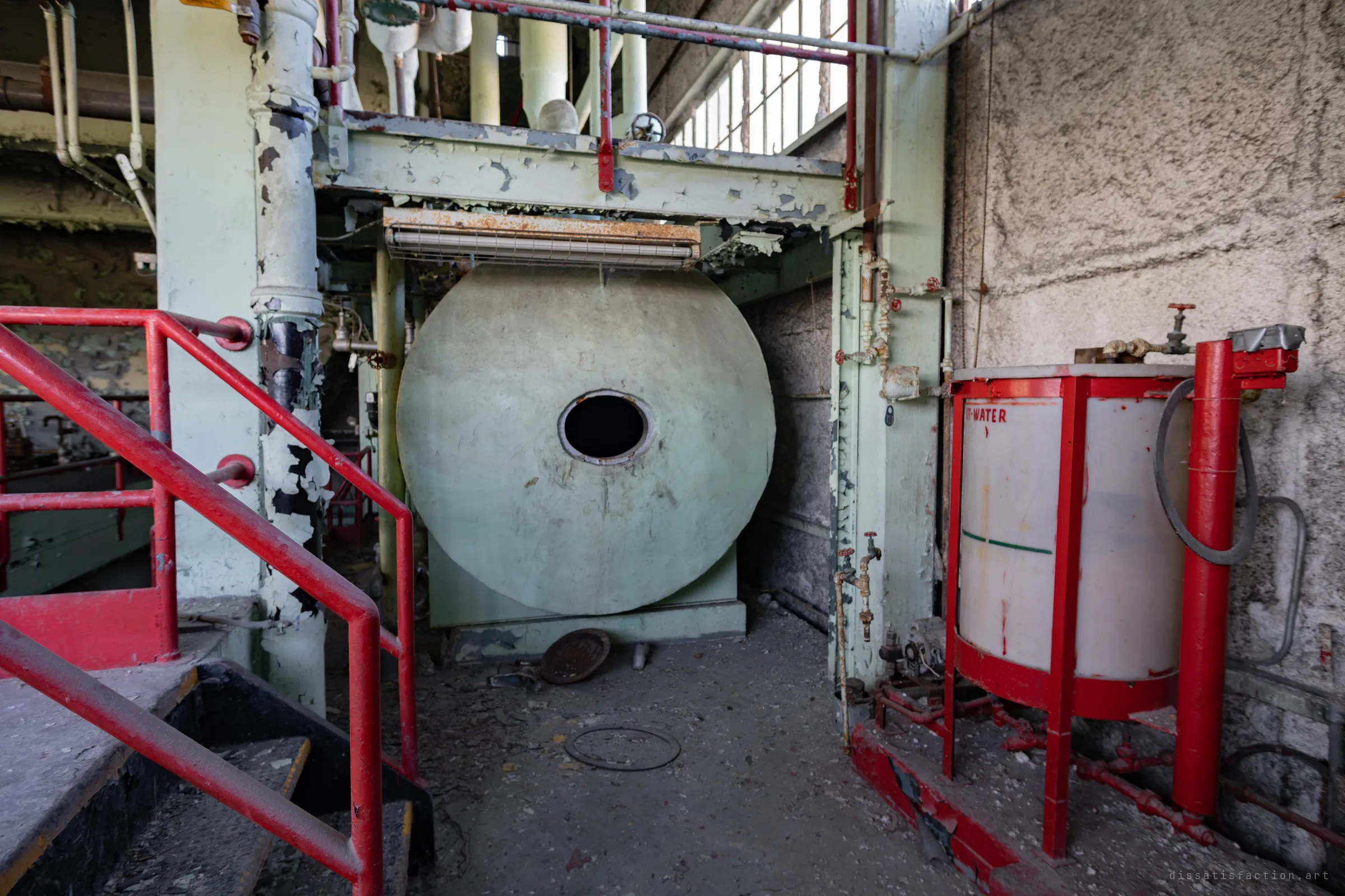 The round, pale green end of a tank with an open manhole, under a steel gantry, beside a red-framed treatment tank and a red-railed stair.
