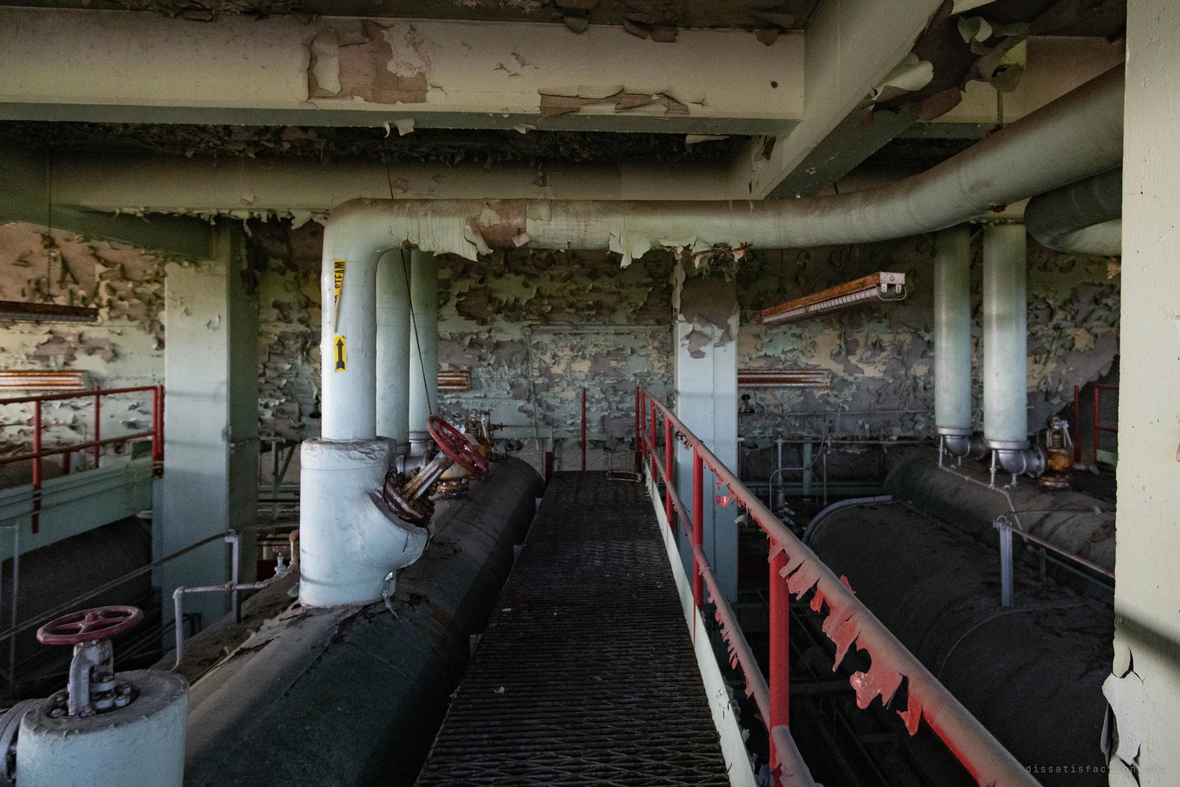 A narrow grated walkway runs between two dark boiler drums, with valves and steam risers on either side and walls blistered with peeling paint.