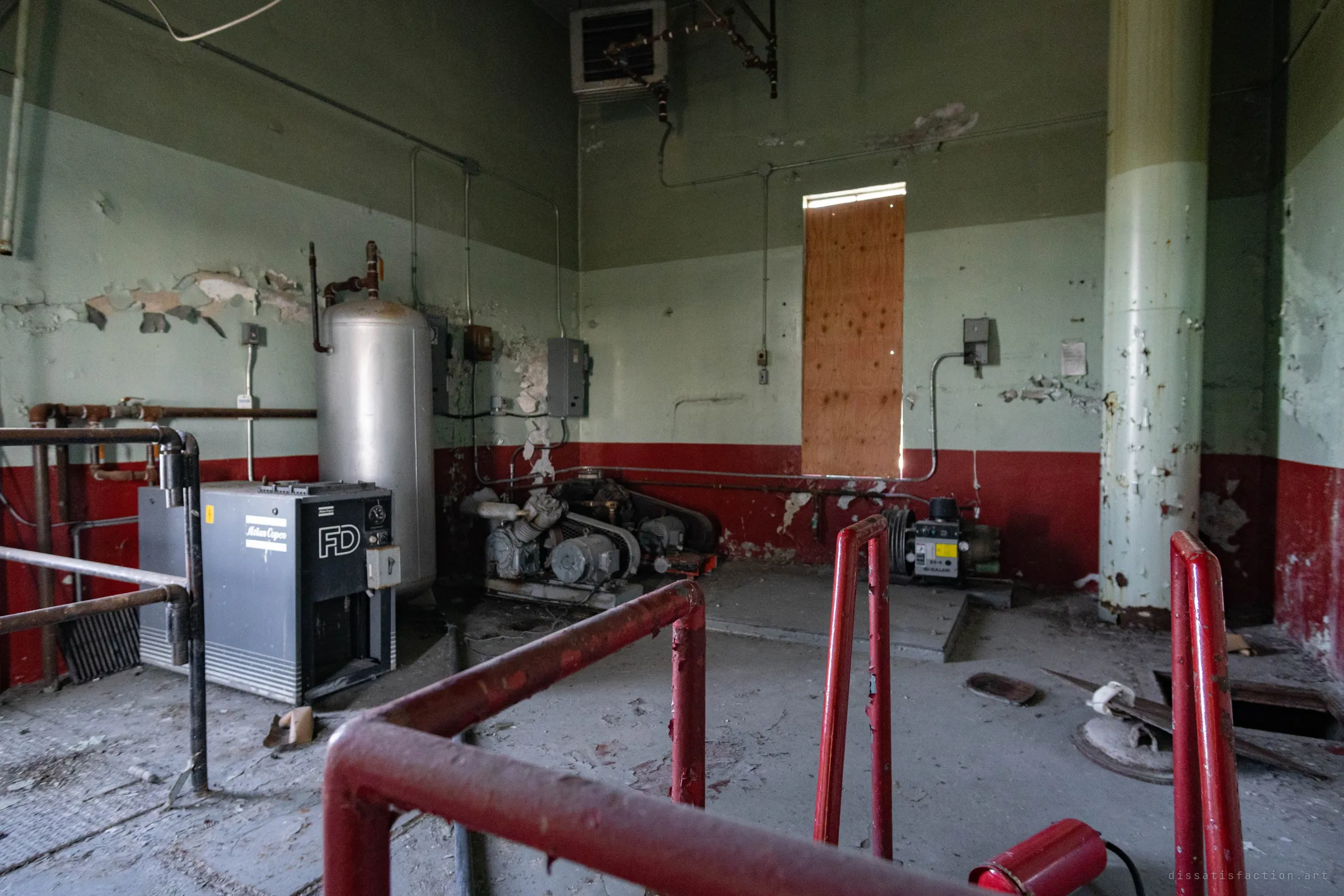 A red-and-green compressor room: an air dryer, a steel receiver tank and belt-driven compressors by a boarded window, seen past red safety rails.