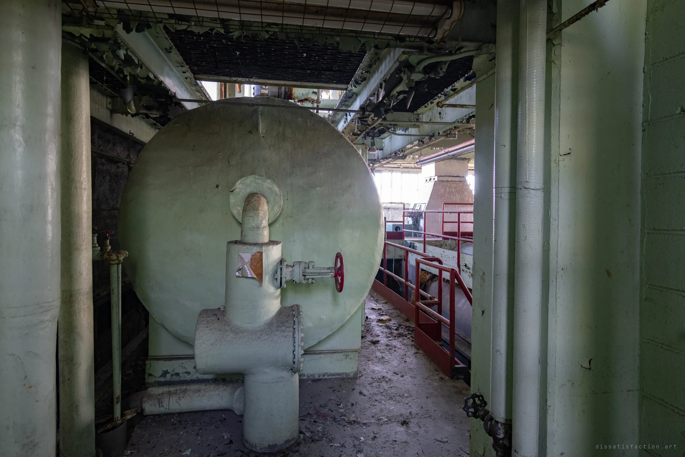The rounded end of a big green tank crowds a narrow passage, a lagged pipe and a red valve on its face, and a red-railed walkway leads toward daylight.