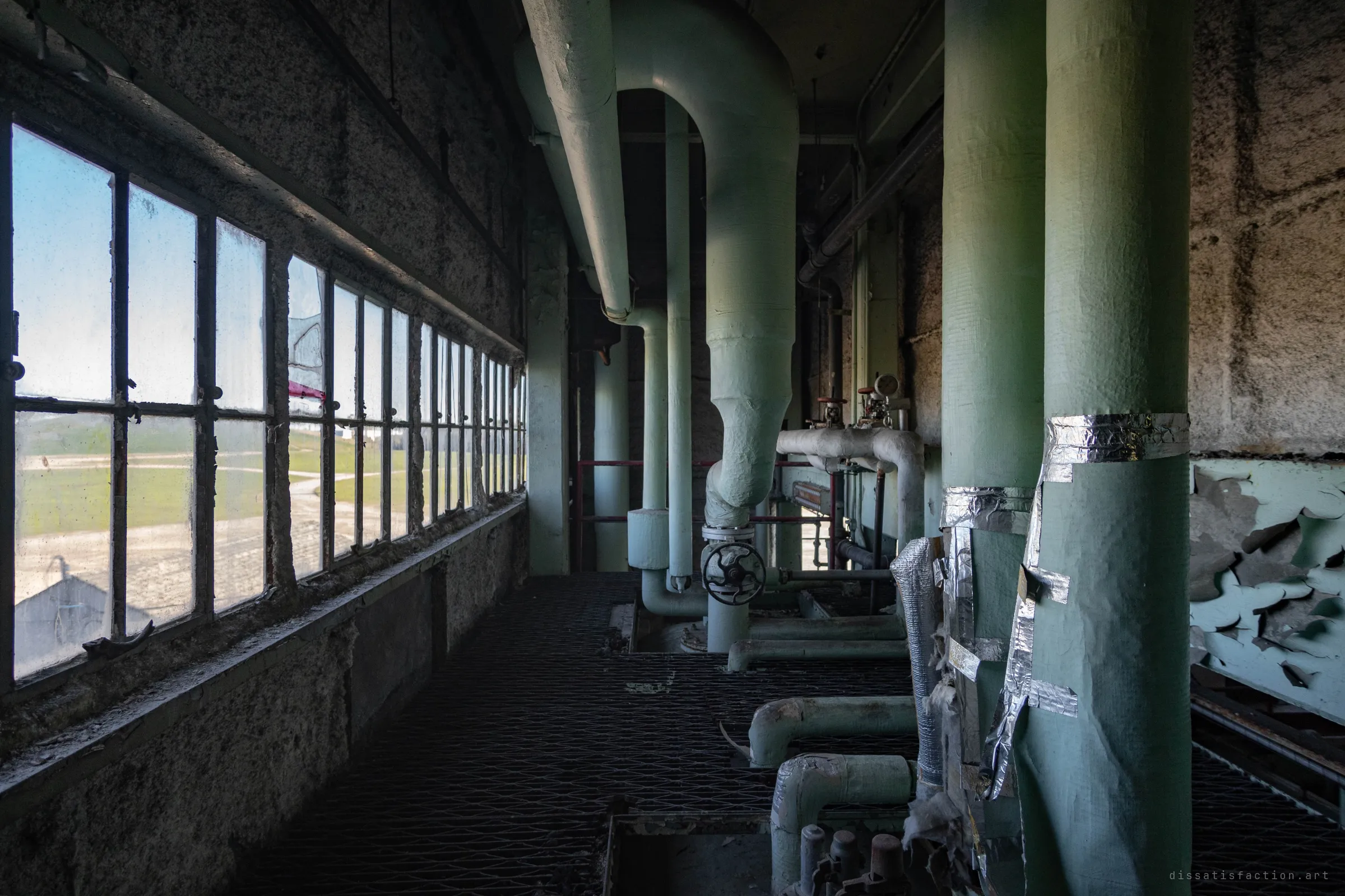 A grated catwalk along a wall of windows, thick green pipes dropping from the ceiling, and flat open land visible through the dirty glass.