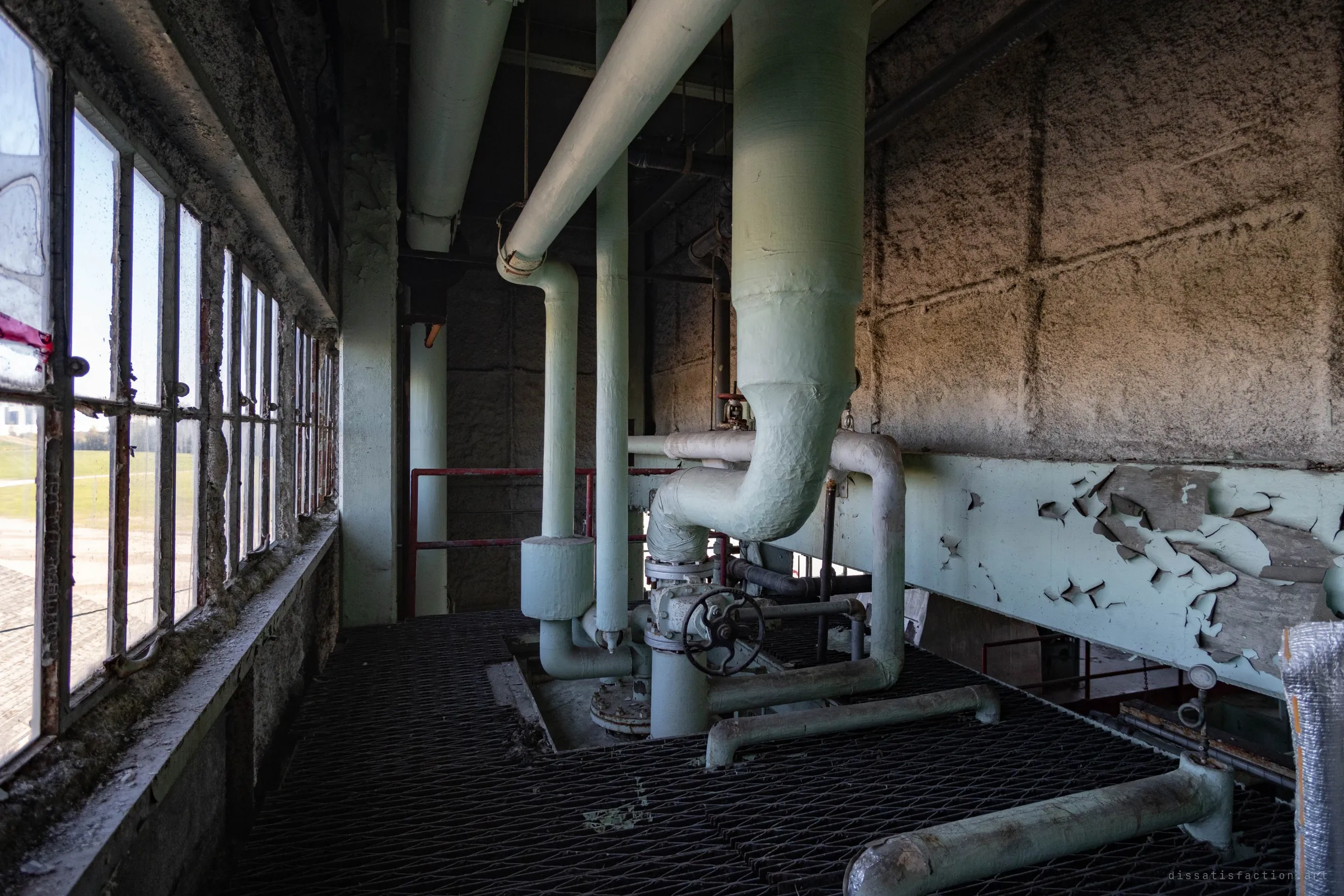 Green steam pipes bend down to a valve on a grated floor beside a wall of windows, the rough concrete wall opposite catching warm light.