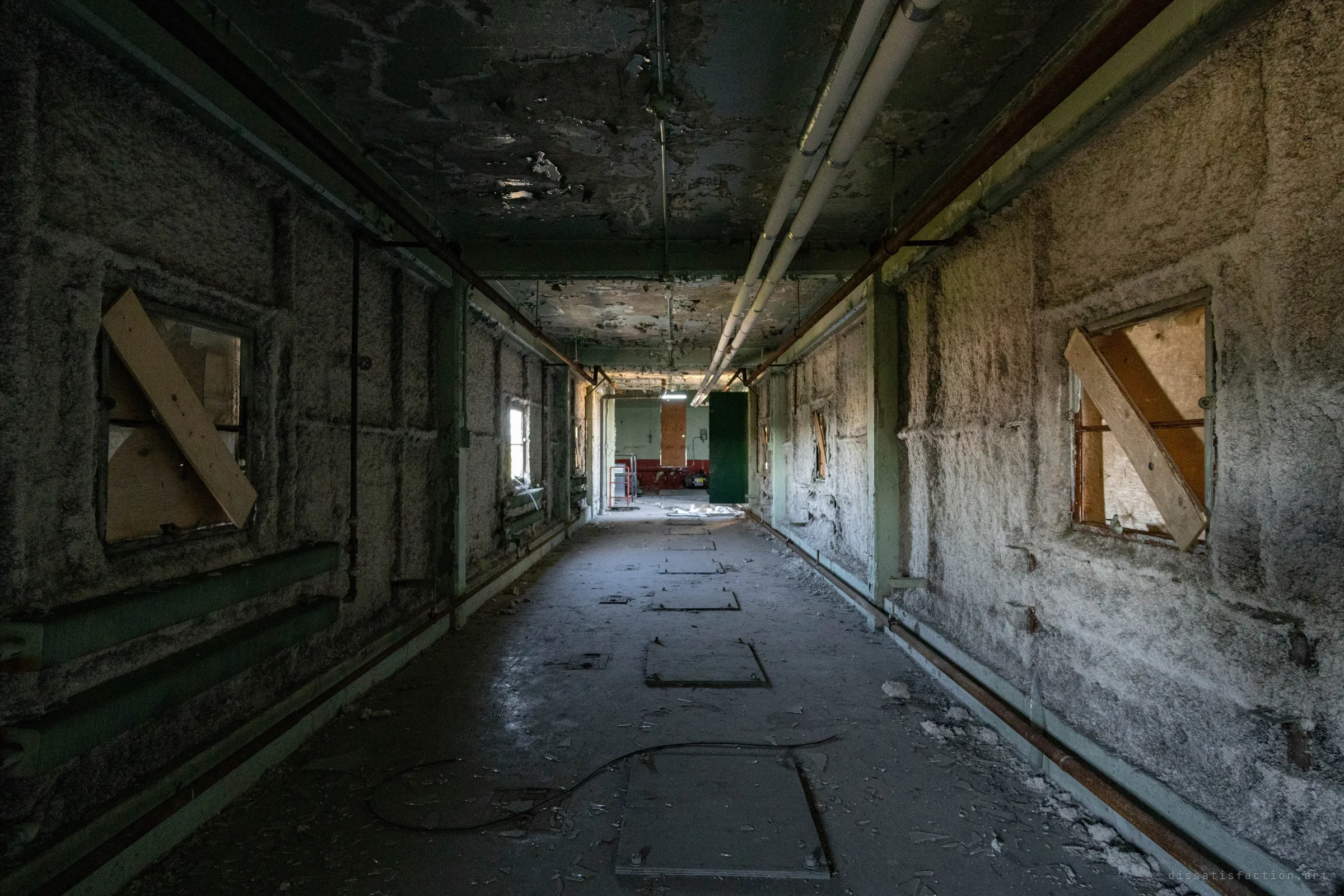 A long corridor lined with rough insulation, its windows boarded with crossed planks and floor hatches leading to a lit room at the far end.