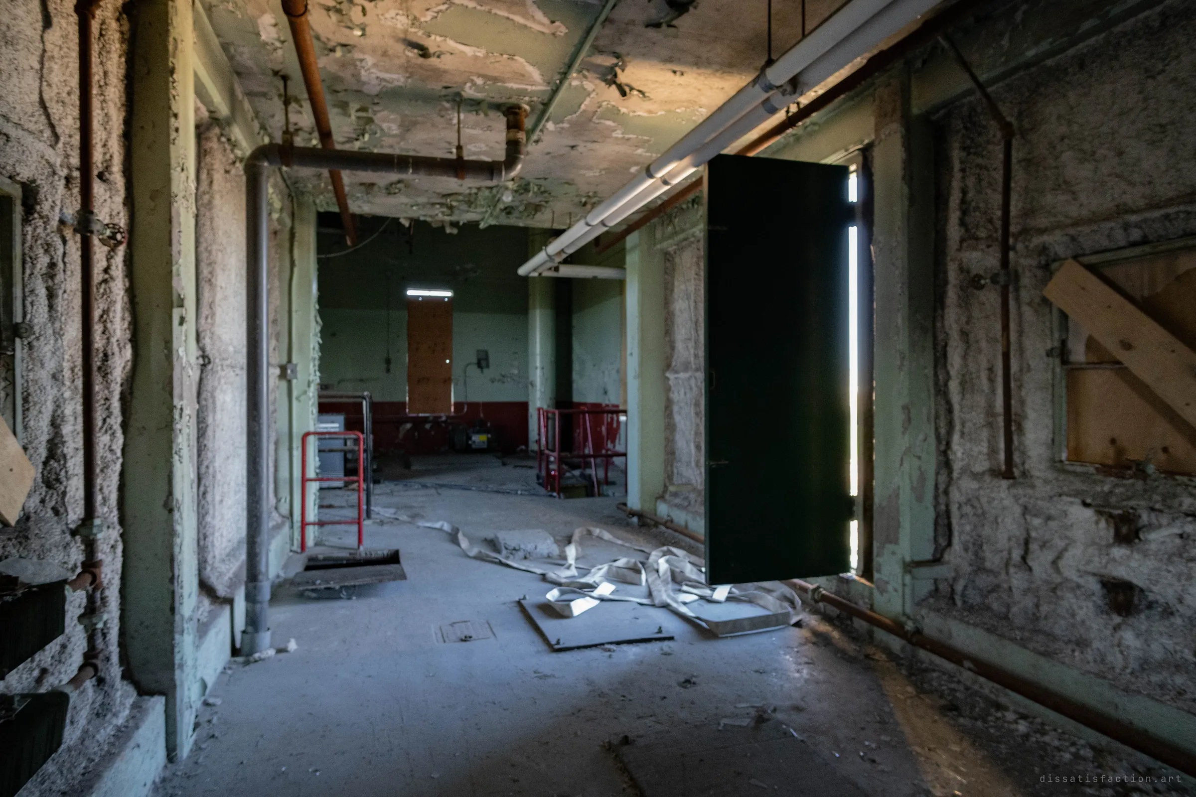 A green door hangs open in a corridor with a fire hose sprawled on the floor, leading on to a red-and-green room with a boarded window.