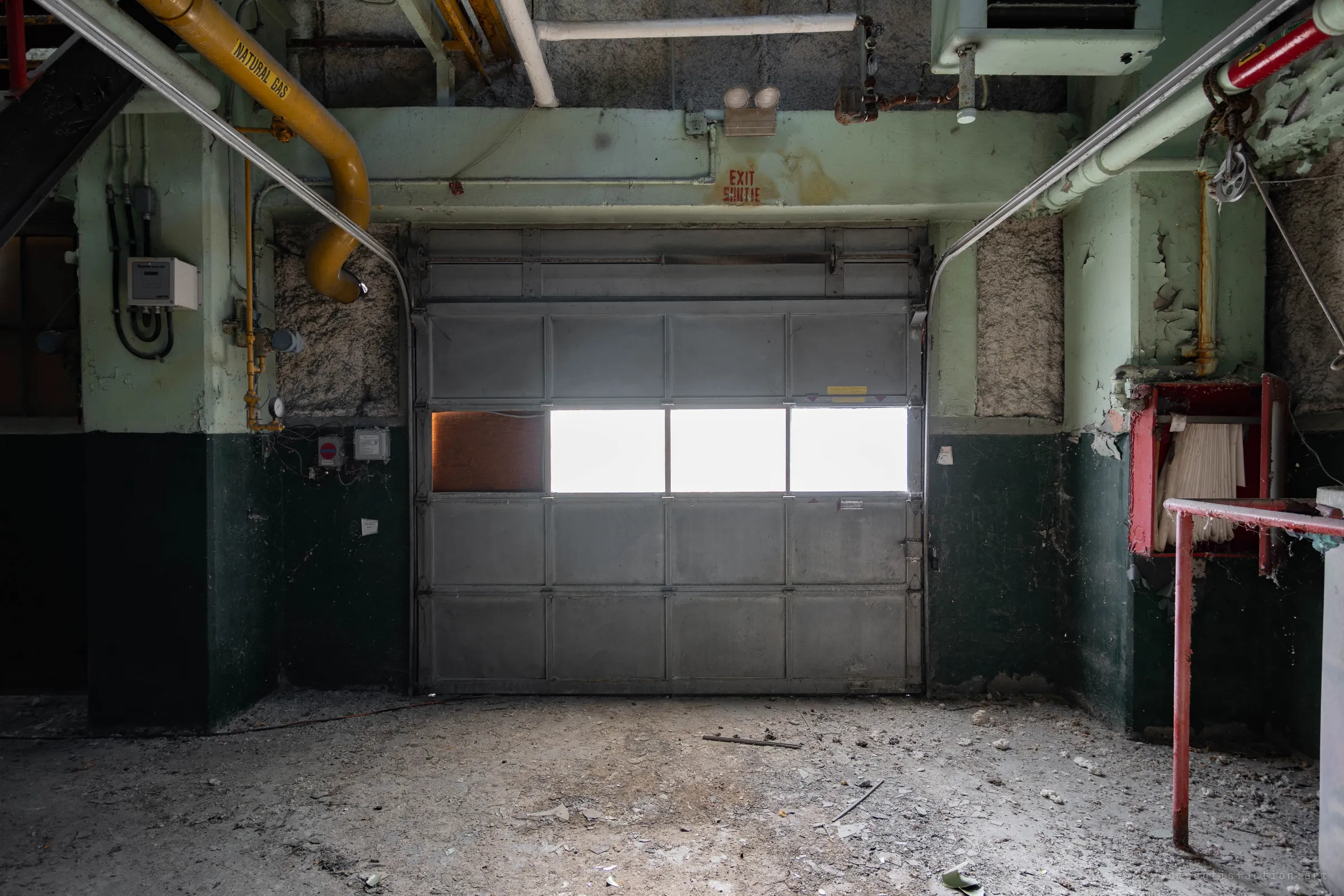 A roll-up loading door with a strip of blown-out windows under a stencilled exit mark, a gas line bending down on the left and a hose cabinet on the right.