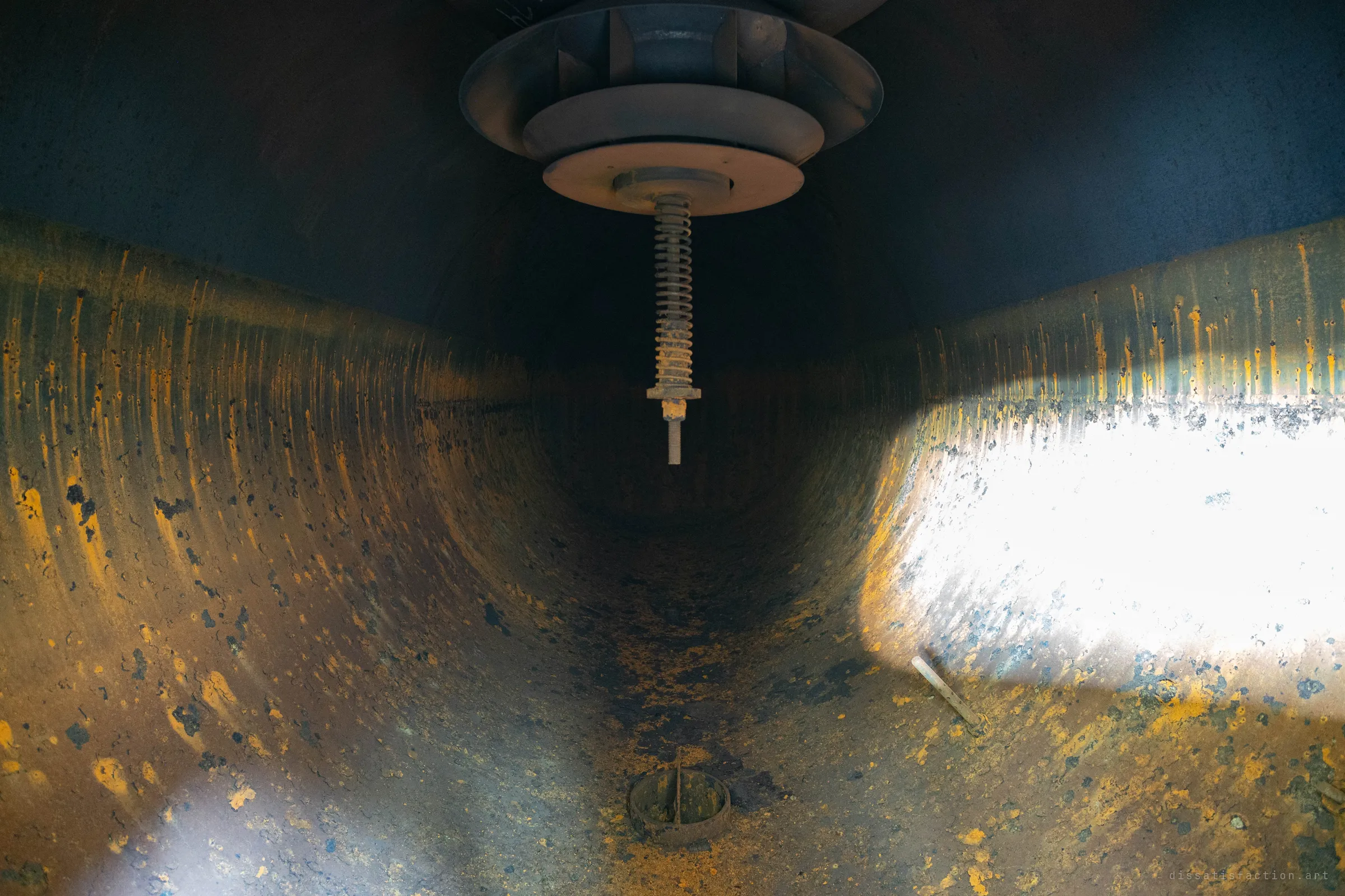 Inside an empty boiler shell: rust-streaked curved walls, a spring-loaded fitting hanging from the top and a patch of hard light on the right.