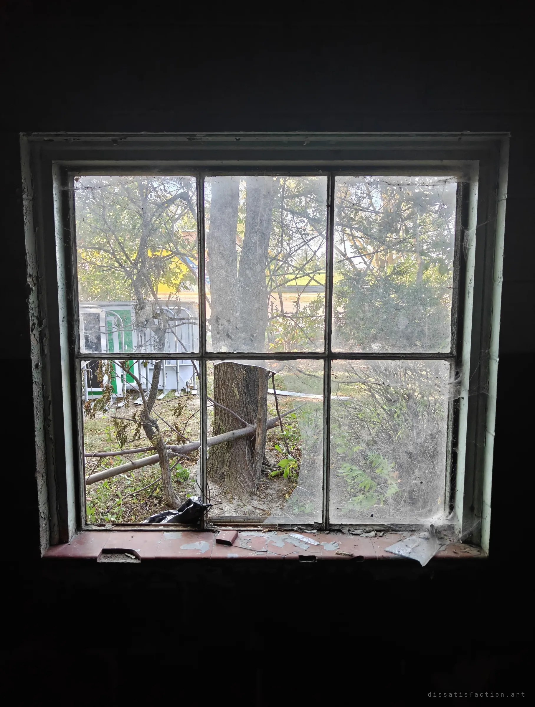 From inside the dark building, a cobwebbed six-pane window with a broken pane looks out on trees and stacked metal barriers. The peeling red sill is scattered with debris.