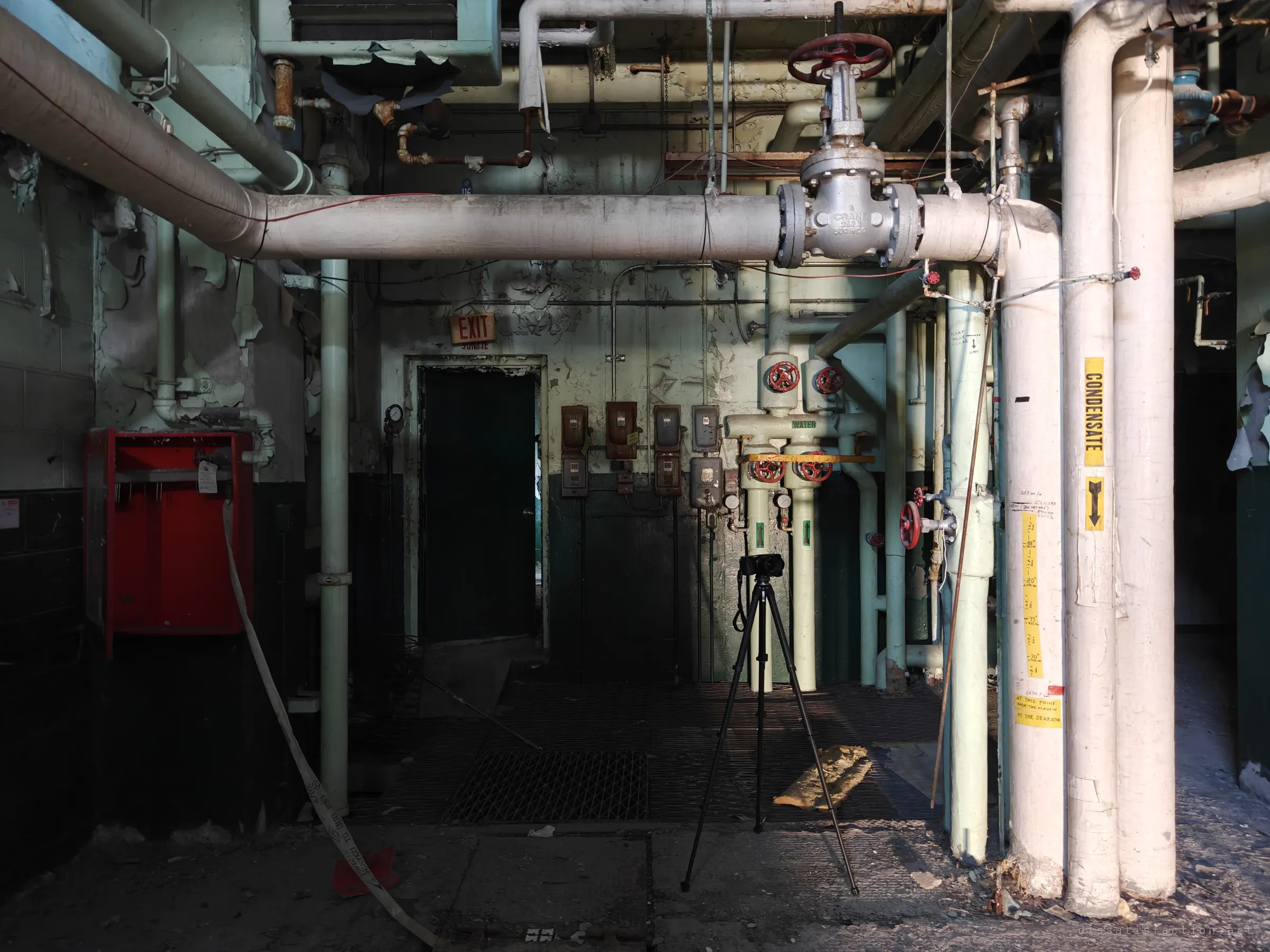 Inside the plant, insulated steam pipes and a large gate valve cross above a grated floor, with gauges, meters and a condensate line marked with handwritten level notes. A tripod stands in the middle of the room.