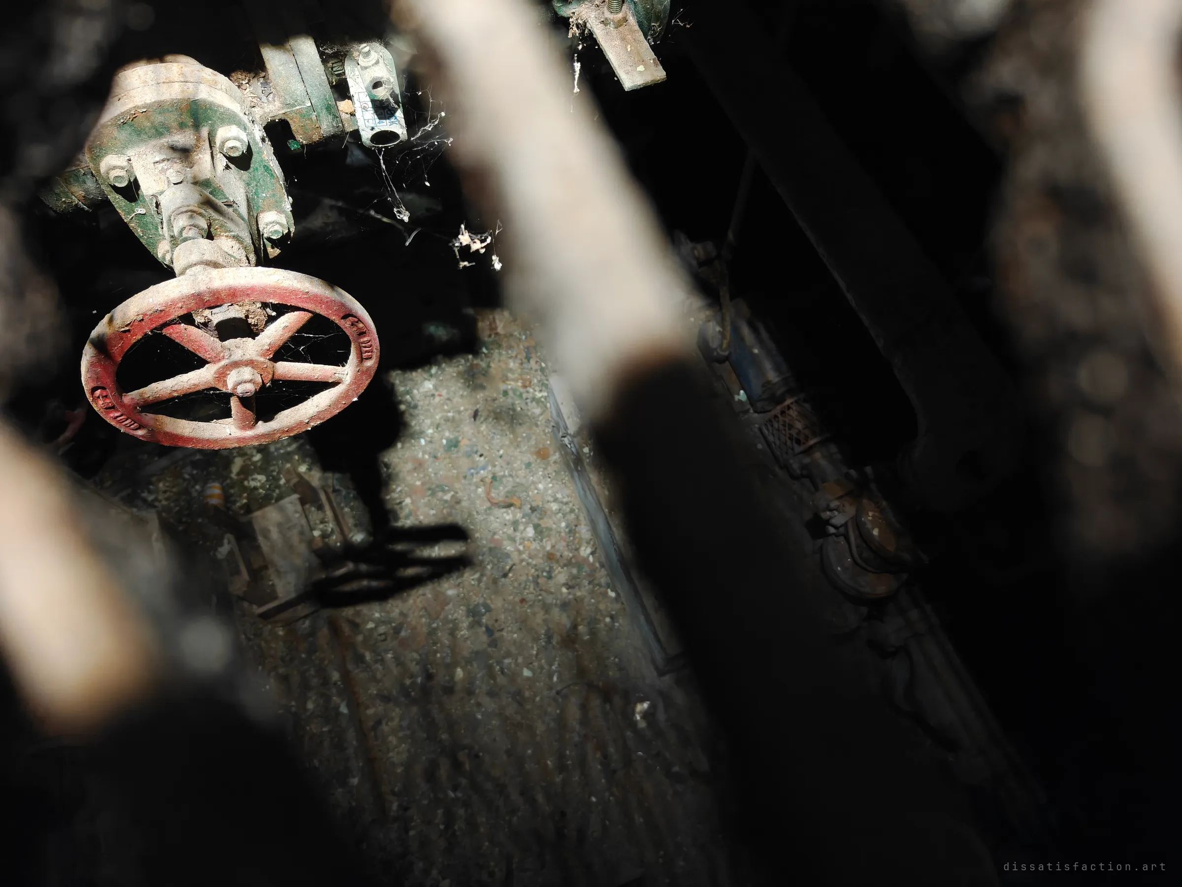 Looking down between blurred pipes to a rust-red valve wheel draped in cobwebs, lit by a shaft of light in a dusty pit. Details left untouched since the plant went quiet.