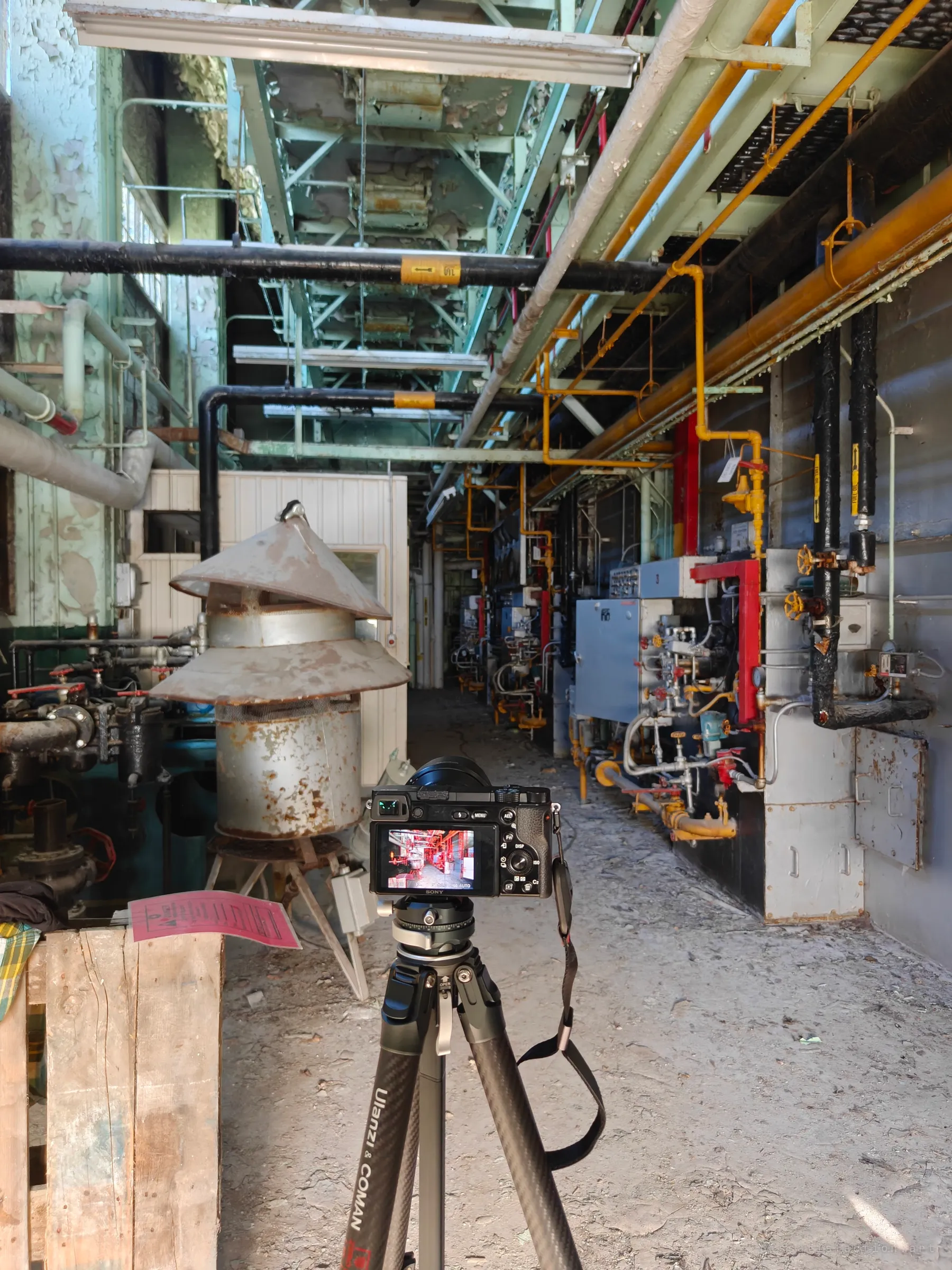 Behind the scenes: a mirrorless camera on a carbon tripod faces down a long row of gas-fired boilers, its screen previewing the red-lit shot. Shows the working setup inside the plant's firing aisle.