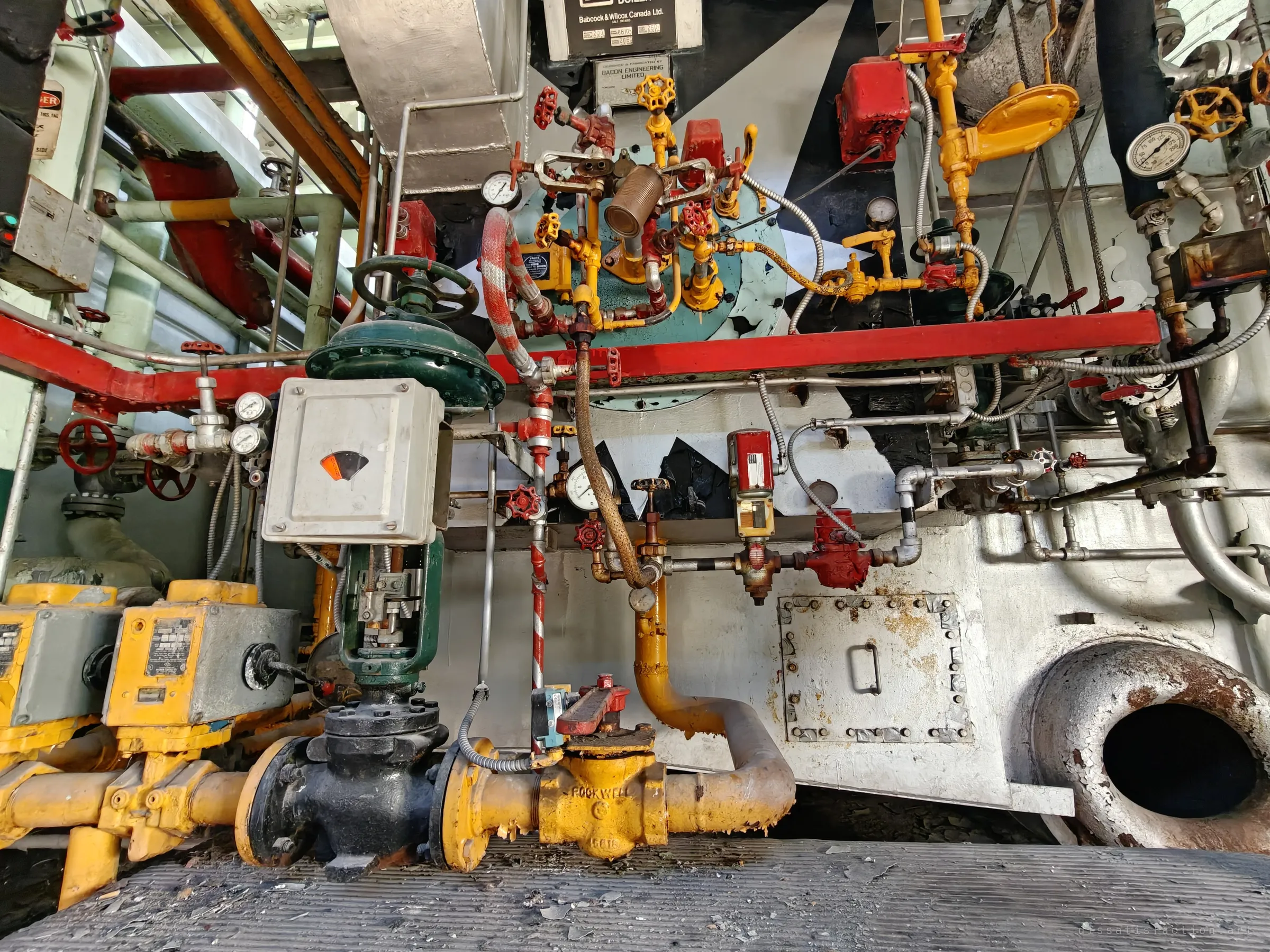 Low wide view of a boiler burner front painted in bold black-and-white chevrons, bristling with red valves, yellow gas piping and pressure gauges above a round inspection port. The colour-coded pipework makes the machinery read almost like a poster.