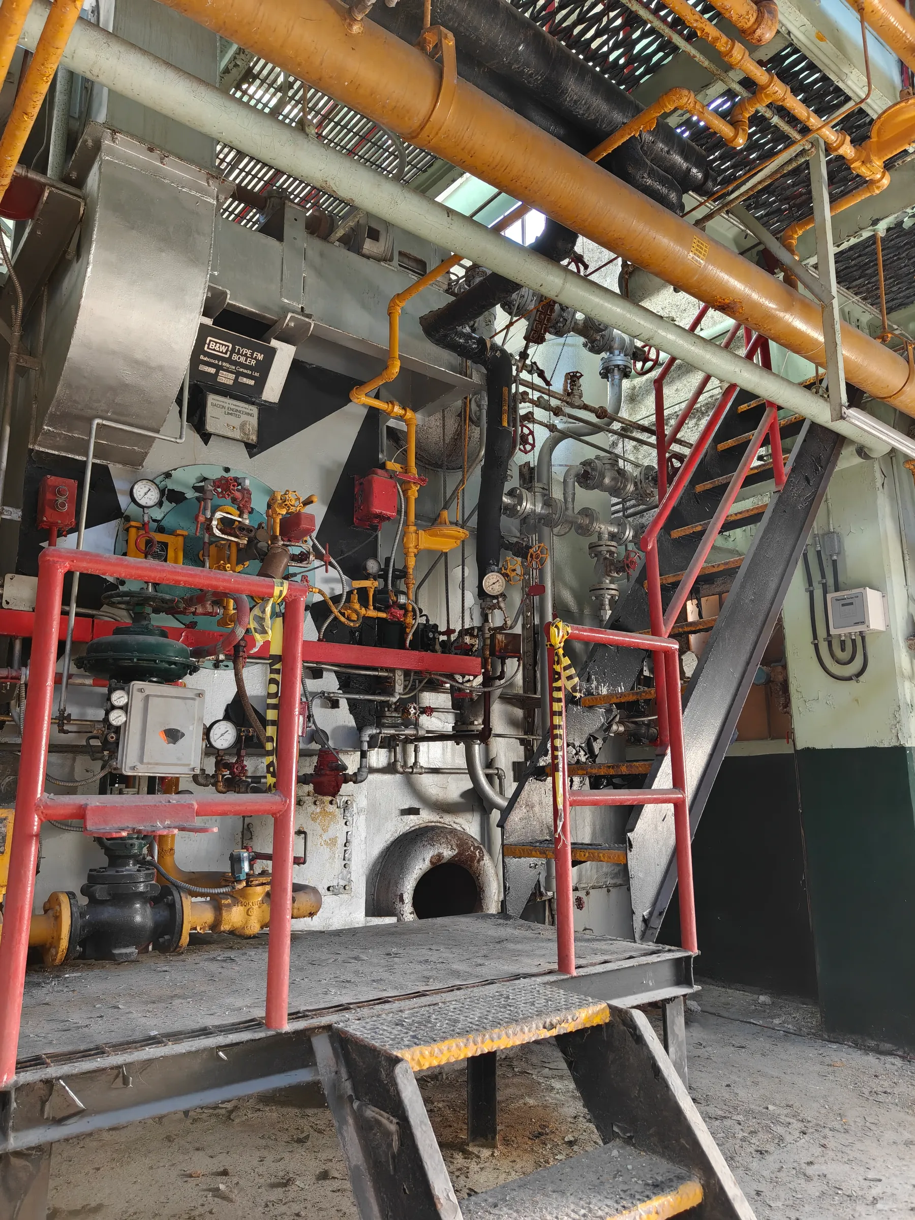 Steel stairs climb to a red-railed service platform in front of a boiler, with caution tape tied across the handrails and yellow gas lines overhead. Gives a sense of the scale and layered access around each unit.
