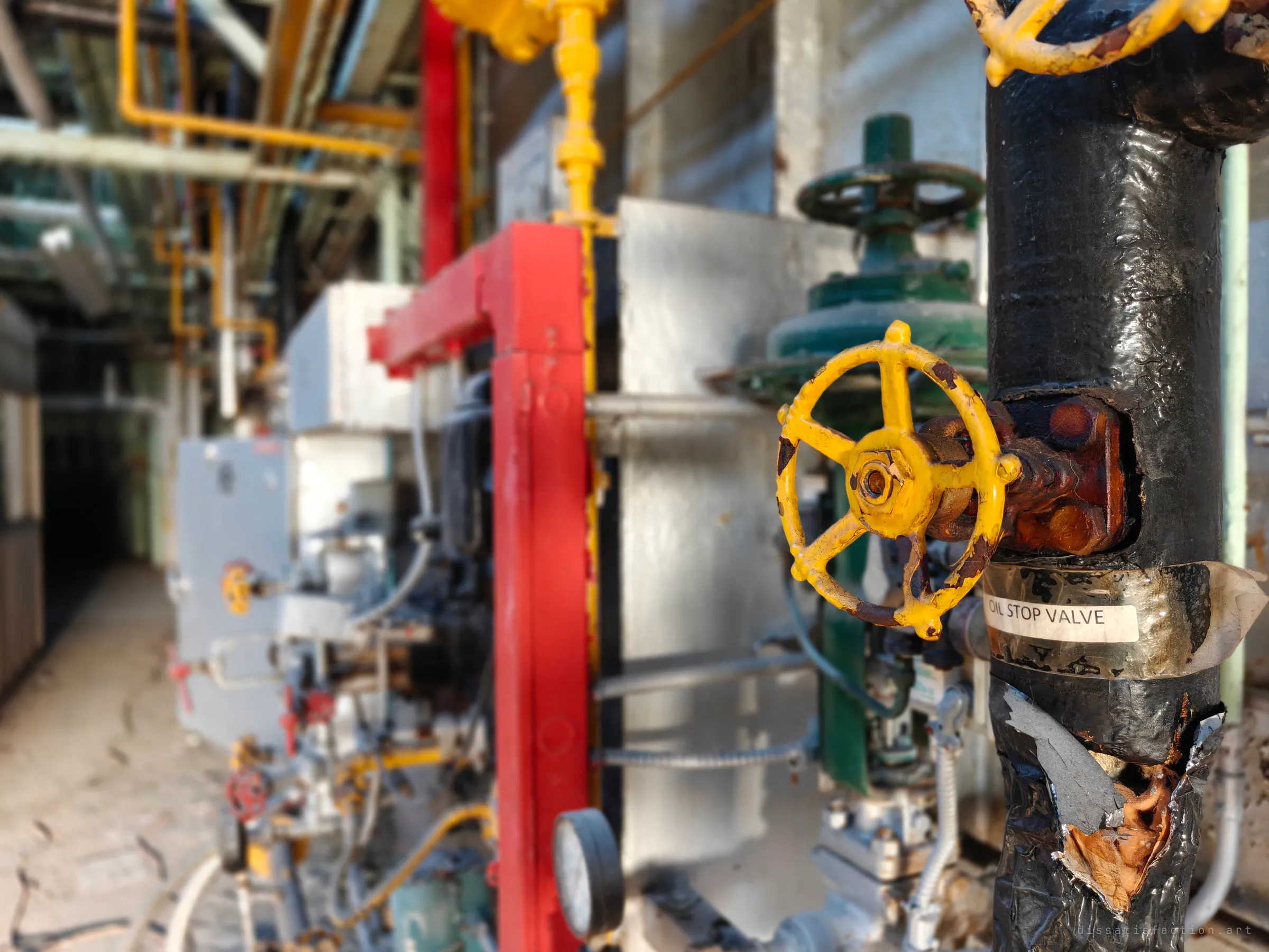 Close focus on a chipped yellow handwheel on a black insulated pipe labelled as an oil stop valve, with the boiler row blurred behind. A small detail of the plant's dual gas and oil firing.
