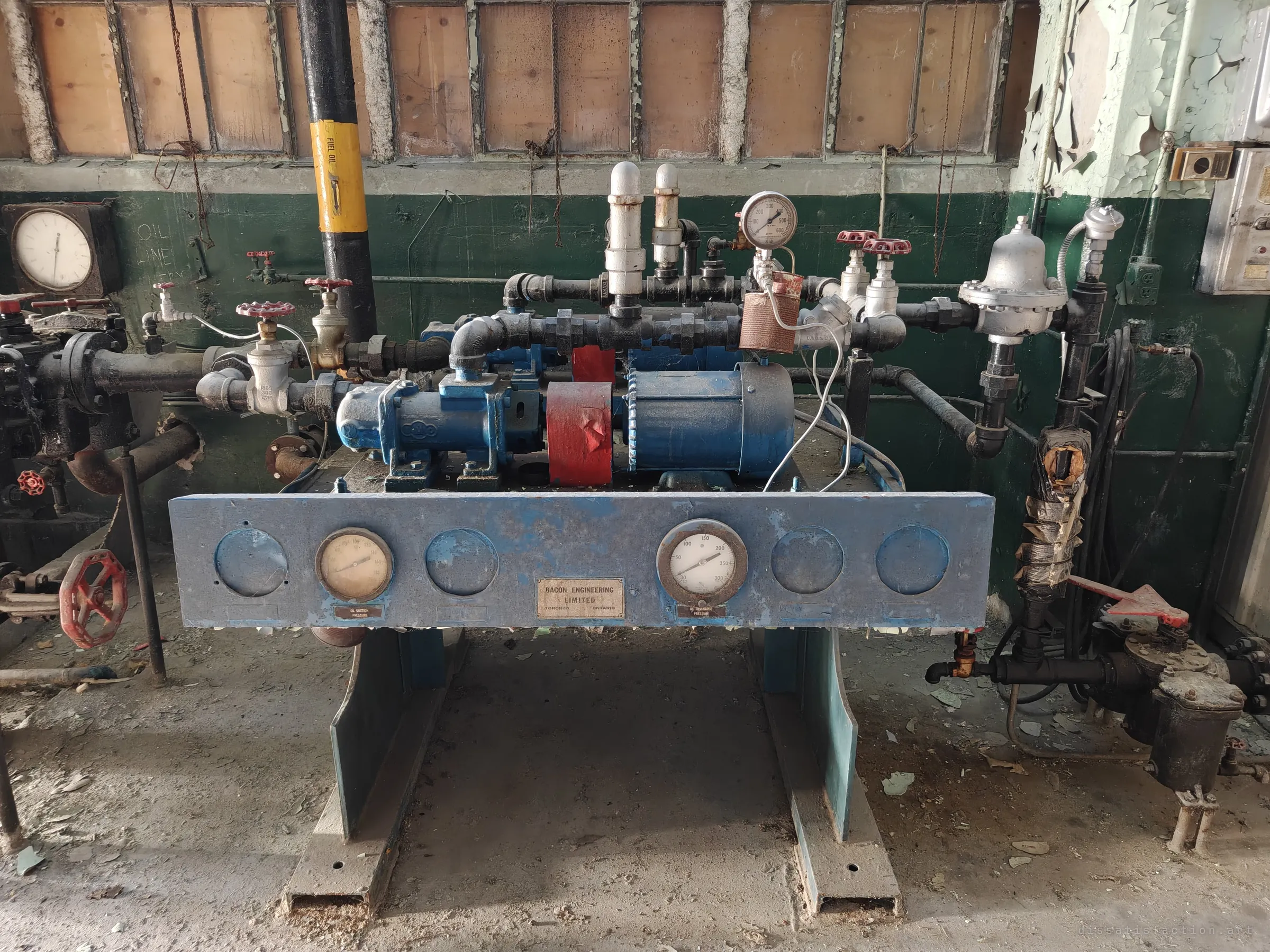 A blue fuel-oil pump skid with twin gauges and a maker's plate sits against a green-and-cream wall below boarded-up windows. Shows the oil supply equipment that fed the burners.