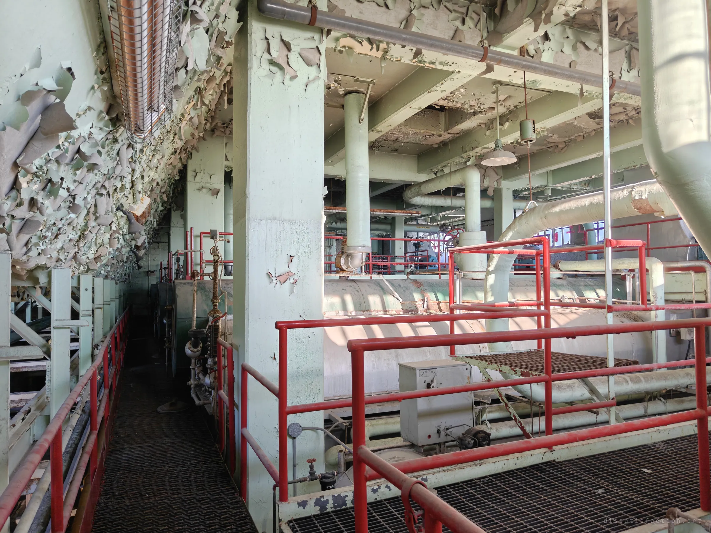 An upper catwalk with red railings runs beside a pale green column, while paint peels from the ceiling in thick curls above horizontal boiler drums. The sunlit decay shows how long the building has been left.