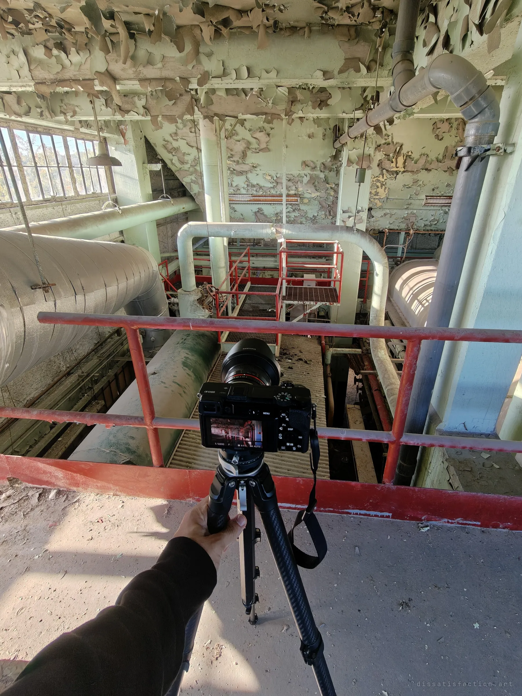 From a red railing on the upper floor, a hand steadies a tripod-mounted camera aimed down into a pit of drums, catwalks and looping insulated pipes under a peeling ceiling. A behind-the-scenes look at shooting from height.
