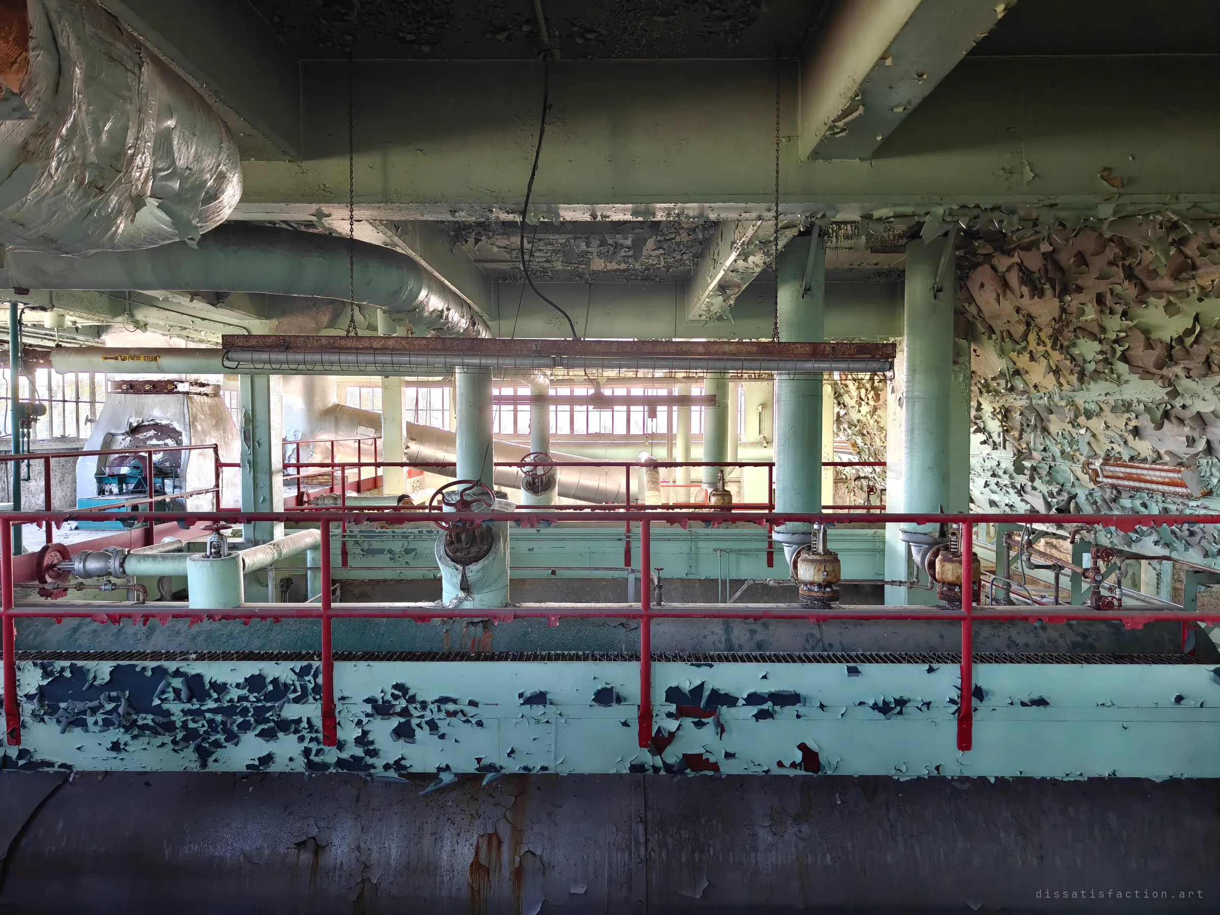 Looking across the top of the boilers: a long red railing, heavy valves and safety valves on the drums, and a wall shedding green paint, with bright industrial windows beyond. The symmetry of the upper deck in one frame.
