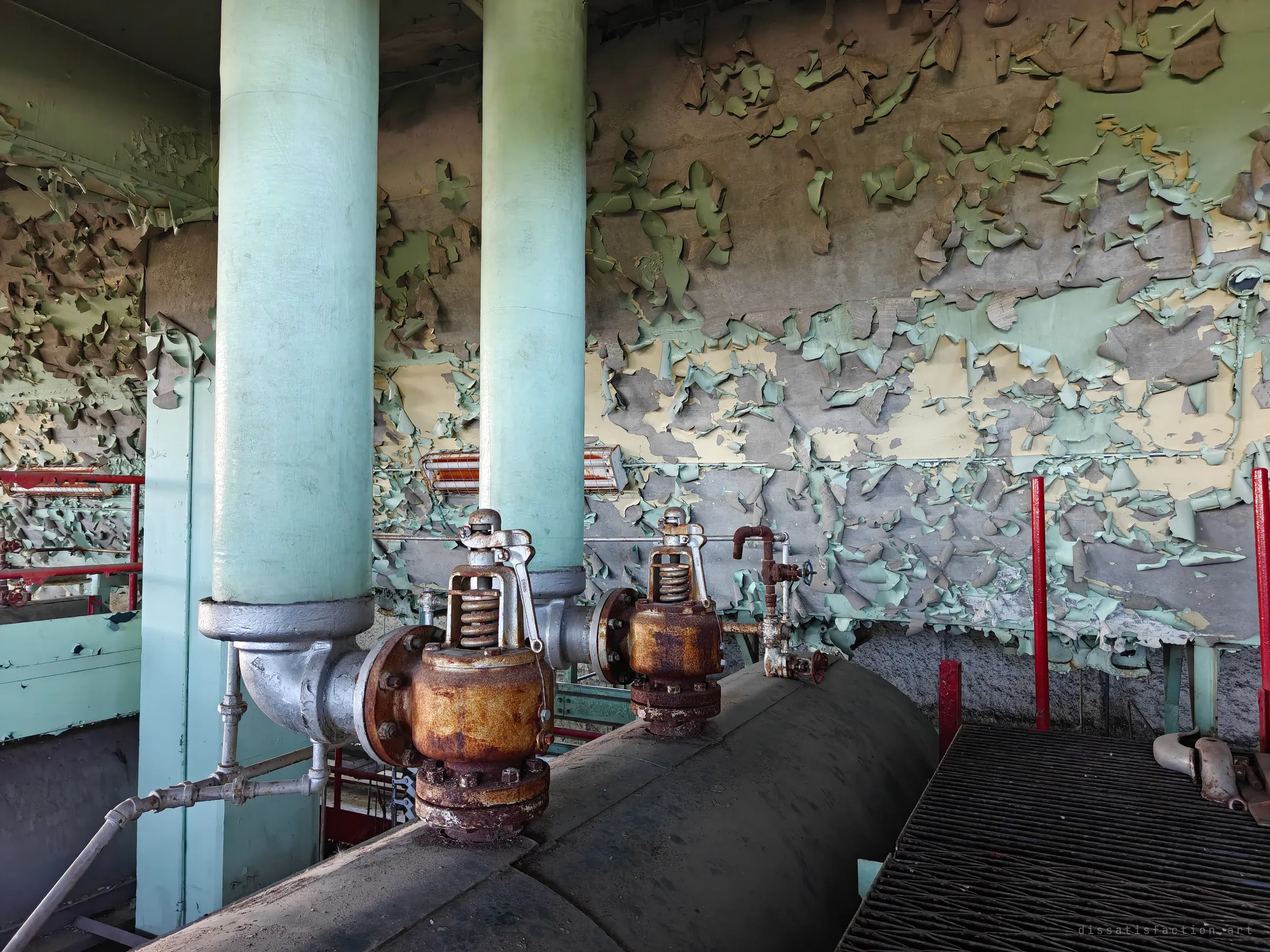 Two rusted spring safety valves sit on a dusty steam drum between pale teal columns, against a wall of curling green and cream paint. A close study of the textures on the plant's top floor.