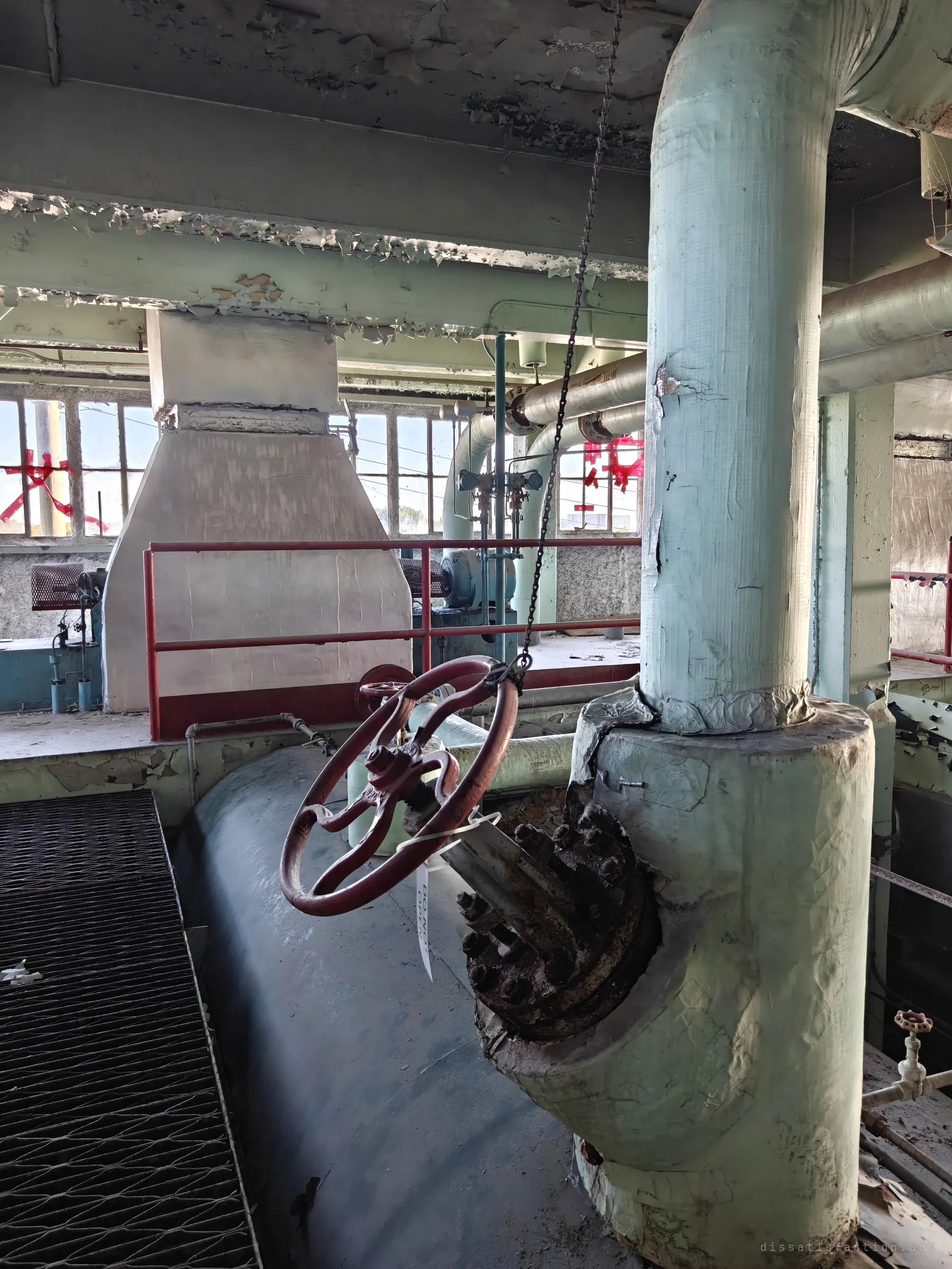 A red valve handwheel hangs from a chain beside a thick insulated steam pipe rising from a drum, with a white fan housing and taped-up windows behind. The dangling chain shows how valves overhead were worked by hand.