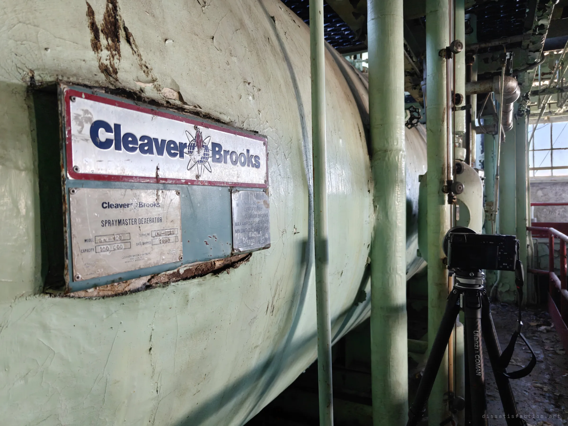 A pale green deaerator tank with its manufacturer's badge and rating plates, a camera on a tripod standing beside it on the catwalk. The maker's plate dates the plant's equipment and shows the scale of the boiler house.