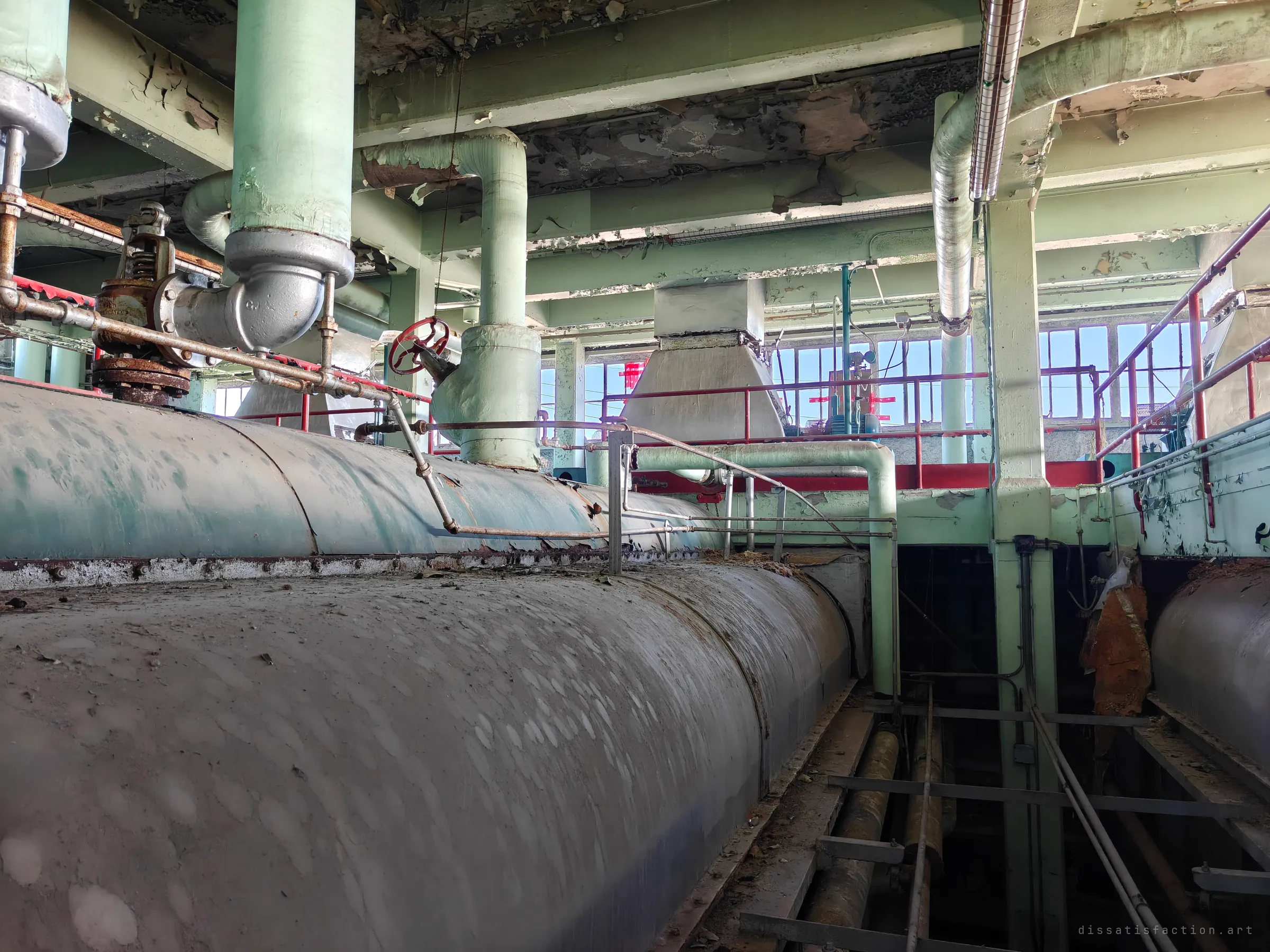 Looking along the tops of two long boiler drums, with insulated pipes, a red valve wheel and red railings under a peeling ceiling. Daylight pours through the tall windows of the upper floor.
